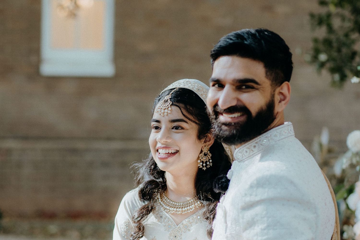 A couple in traditional South Asian wedding attire pose together against a stone wall background.