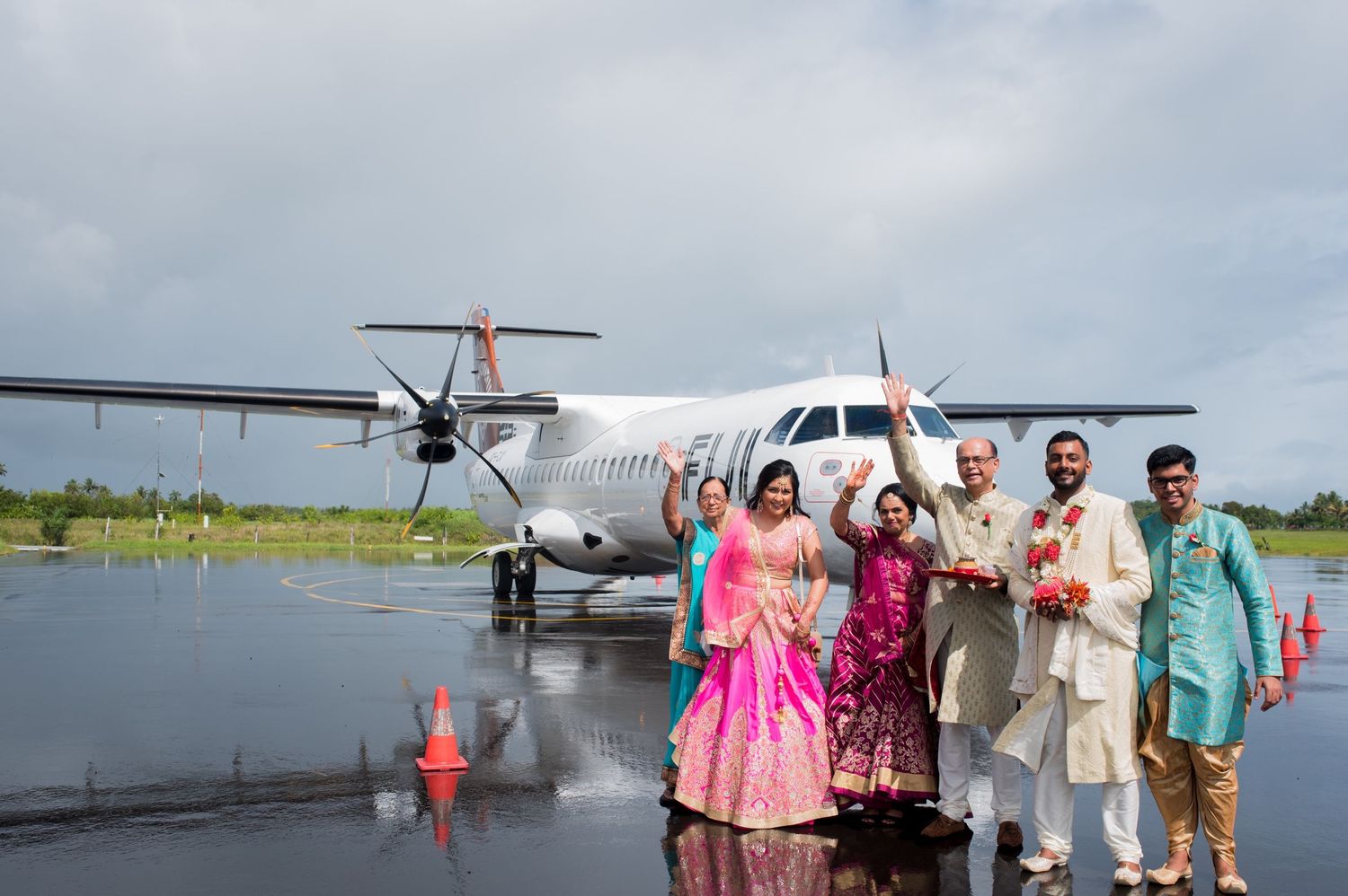 Wedding party poses together in traditional Indian attire on an airport tarmac next to a small passenger aircraft.