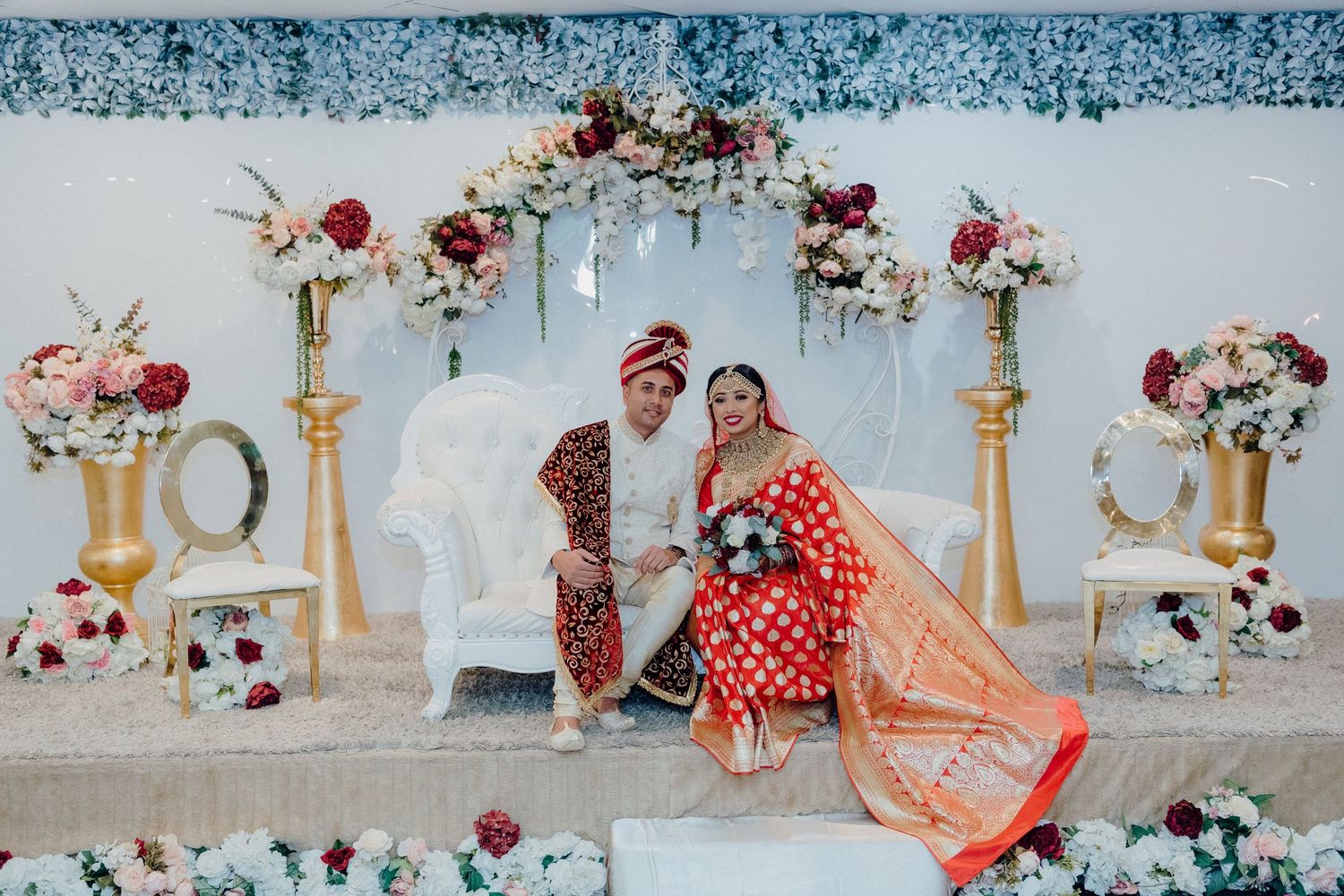 Wedding portrait against white backdrop with gold pillars and pink and red floral arrangements.