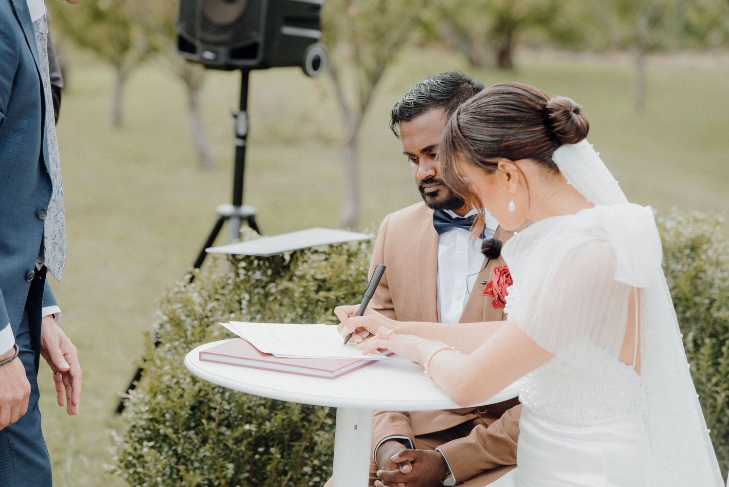 Couple signing marriage documents at an outdoor wedding ceremony with a white table.