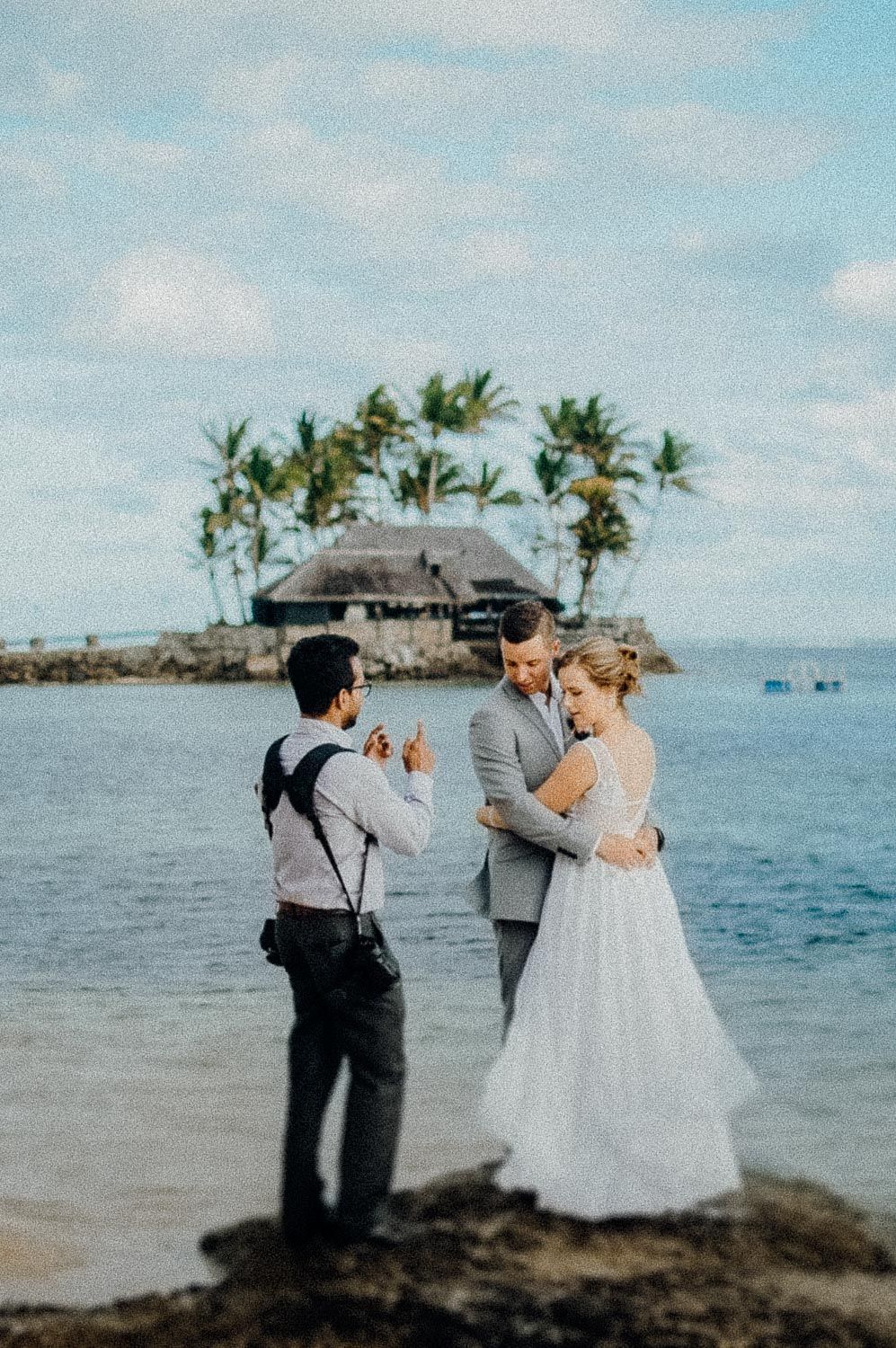 Wedding couple embracing on tropical beach while photographer captures their moment near island.