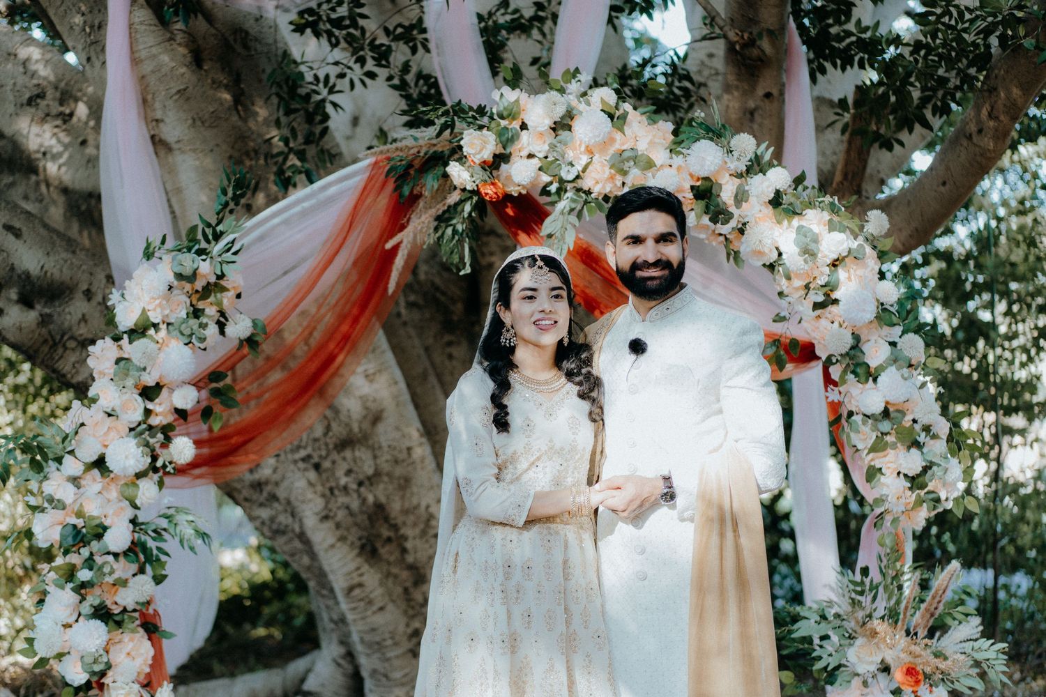 Newlyweds stand under an ornate floral arch decorated with white flowers and coral draping.