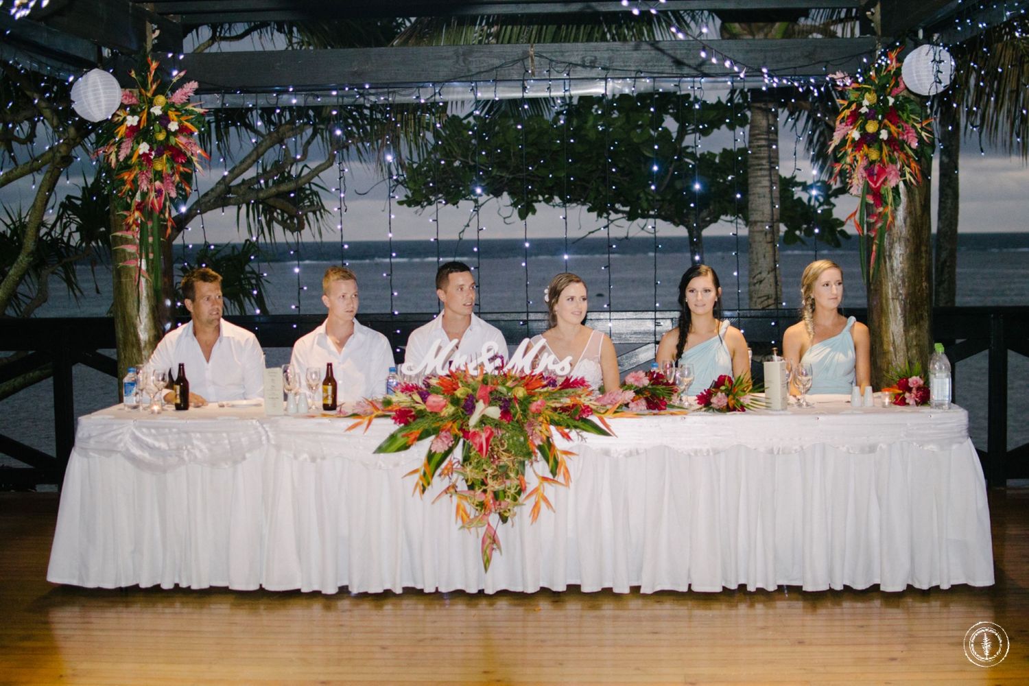 Wedding party seated at decorated head table with tropical floral arrangements.