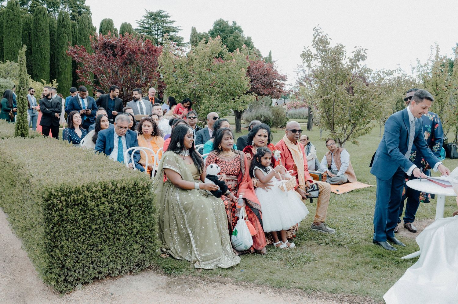 Wedding guests seated on chairs outdoors with cypress trees and colorful foliage in background.