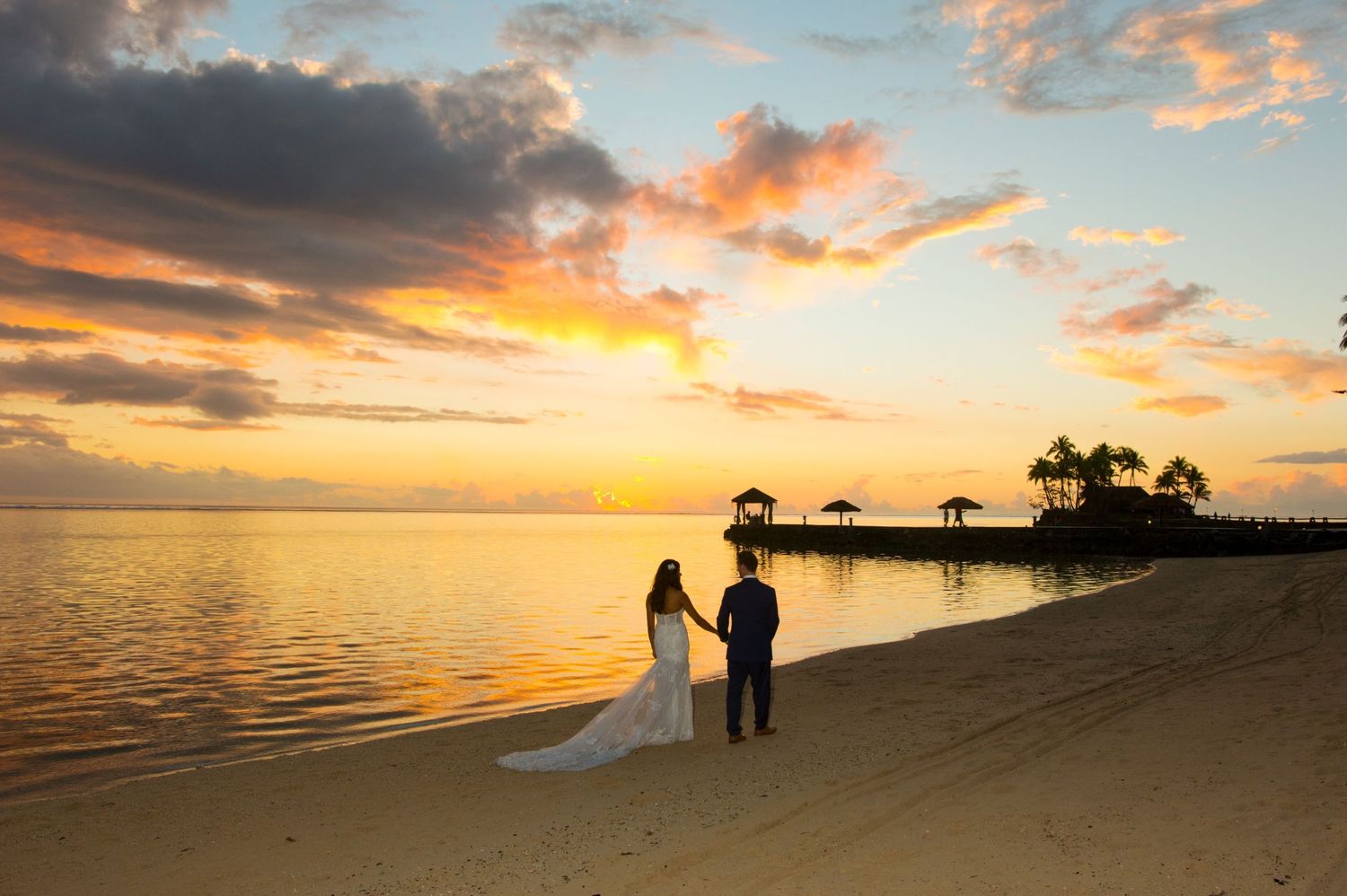 Silhouetted couple standing on a beach during a dramatic orange and gold sunset with palm trees in the distance.