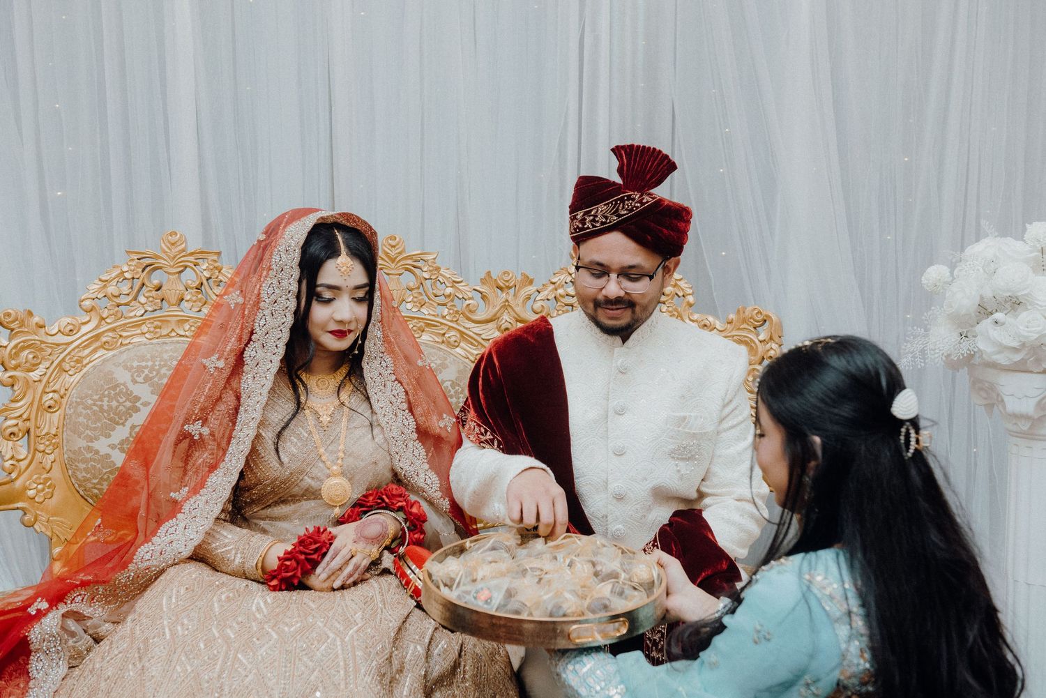 Traditional South Asian wedding ceremony with participants exchanging ritual items on ornate furniture.