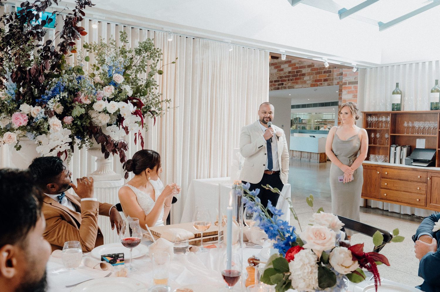 Wedding reception guests enjoying emotional speeches in bright venue with floral decor.