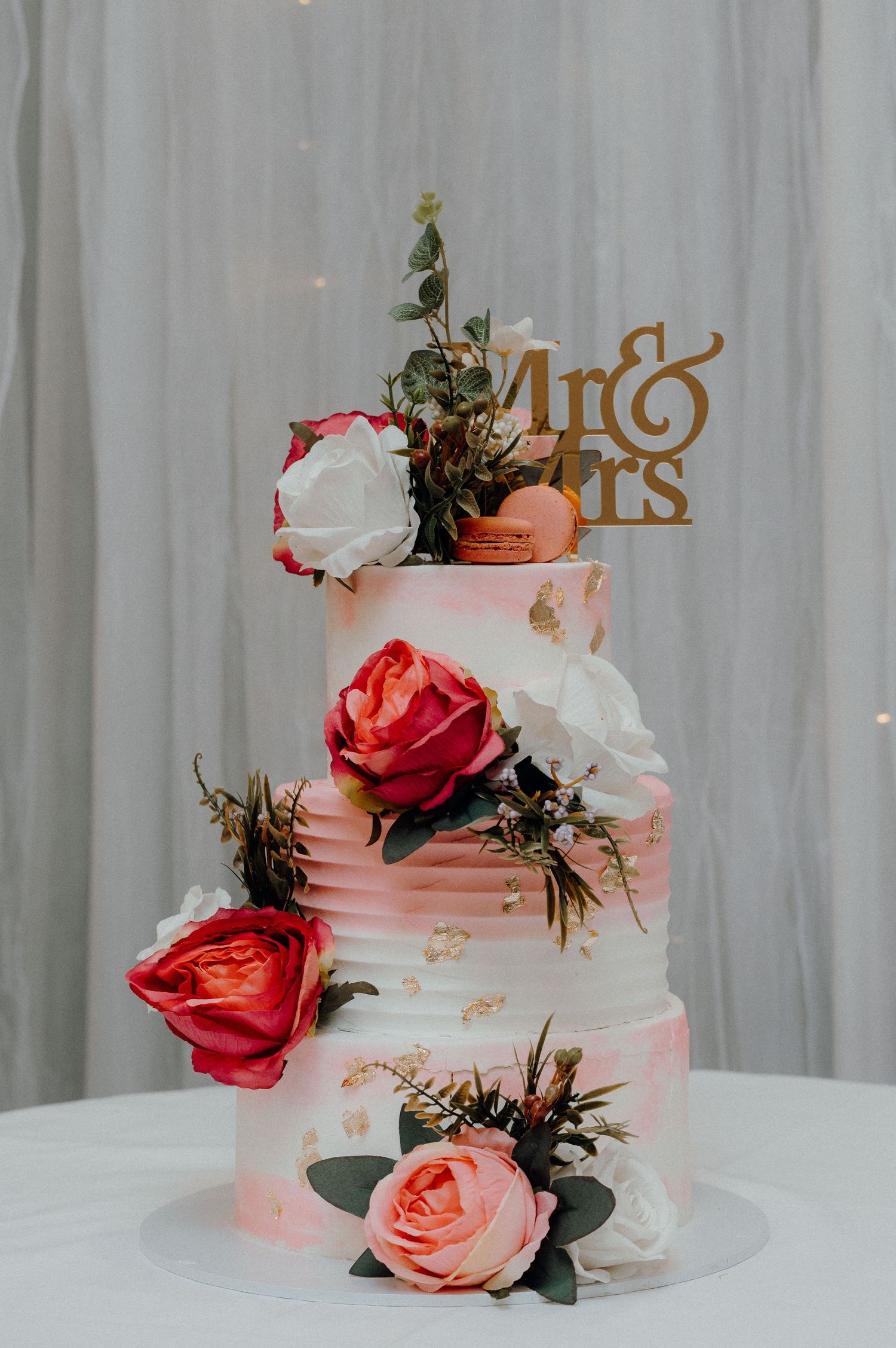 Three-tiered wedding cake decorated with pink and red roses against a white backdrop.