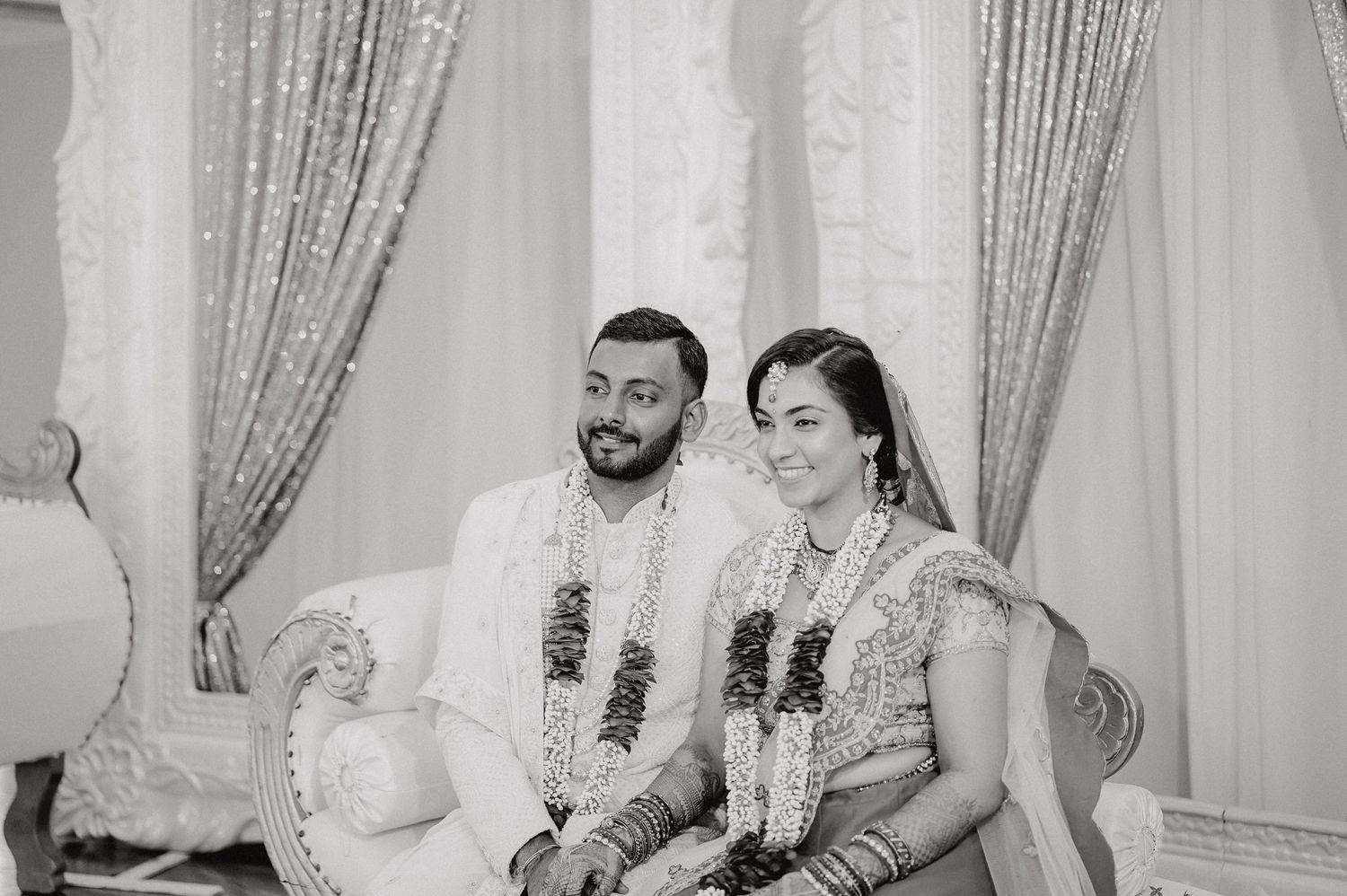 Couple in traditional Indian wedding attire seated on ornate white sofa.