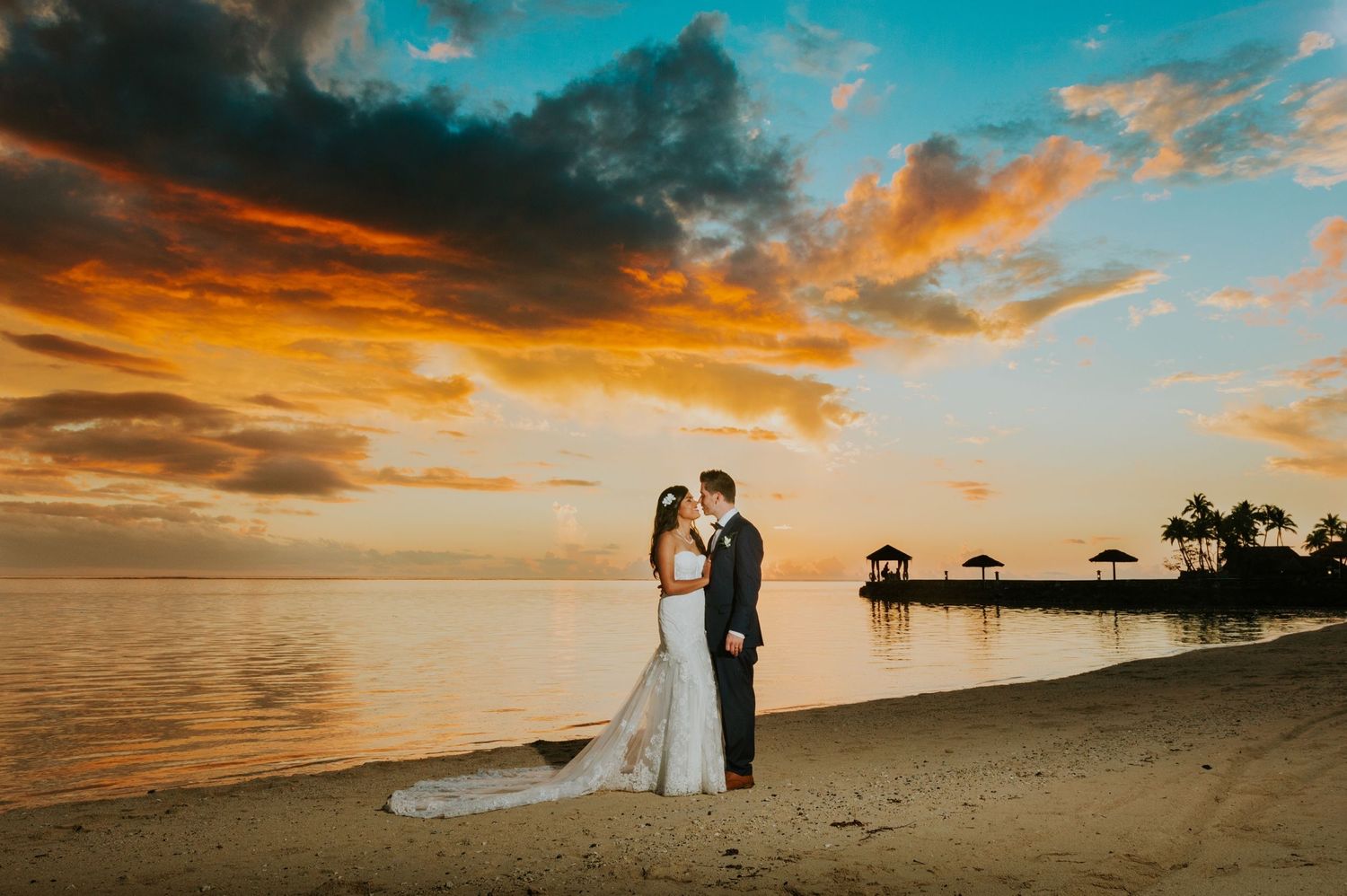 Romantic beach wedding photo at sunset with palm trees silhouetted against orange sky.