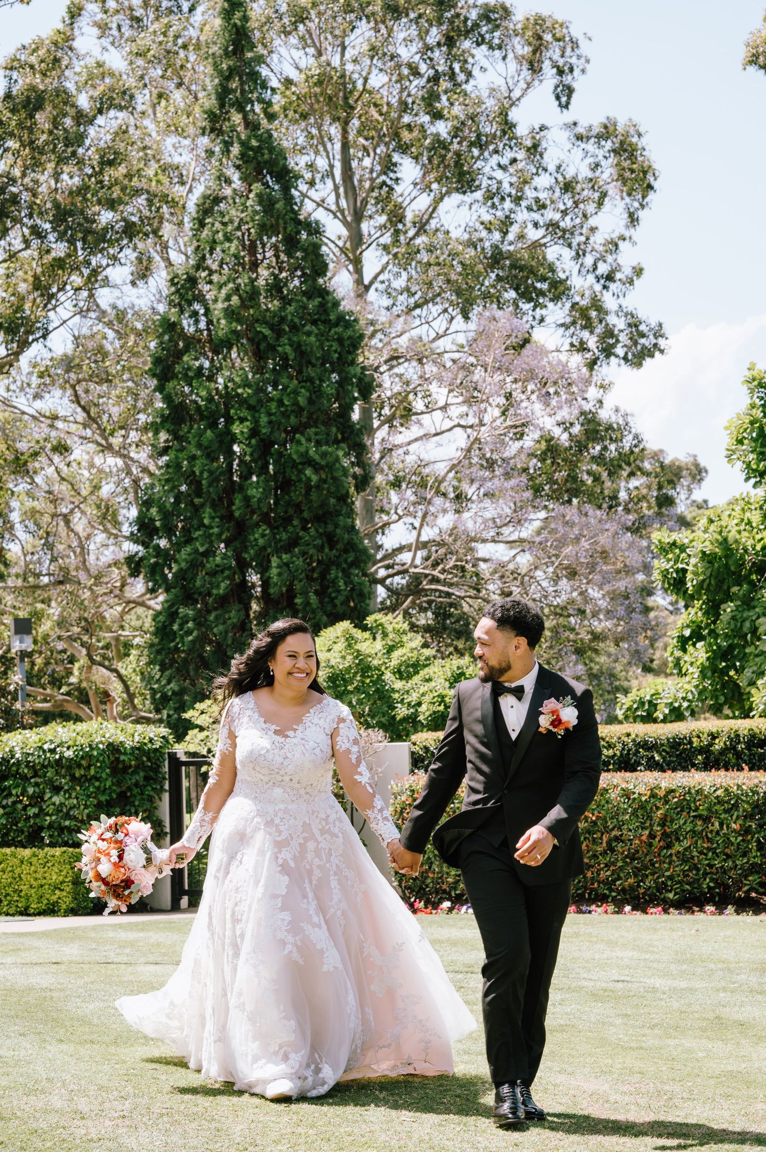 Bride in lace ballgown and groom in black tuxedo walk hand in hand through a sunny garden park.