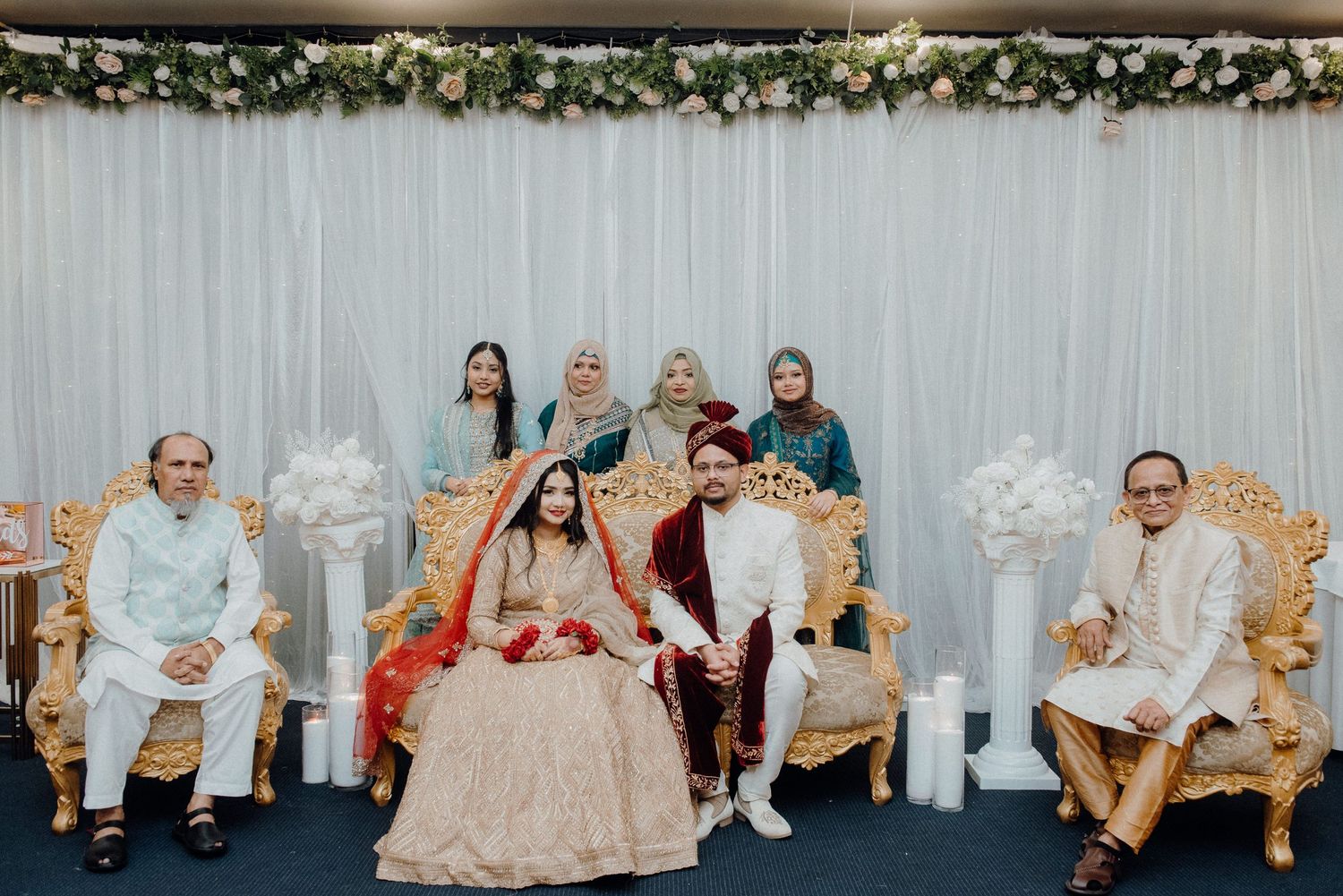Family members seated on ornate chairs at a wedding ceremony with floral decorations and white backdrop.