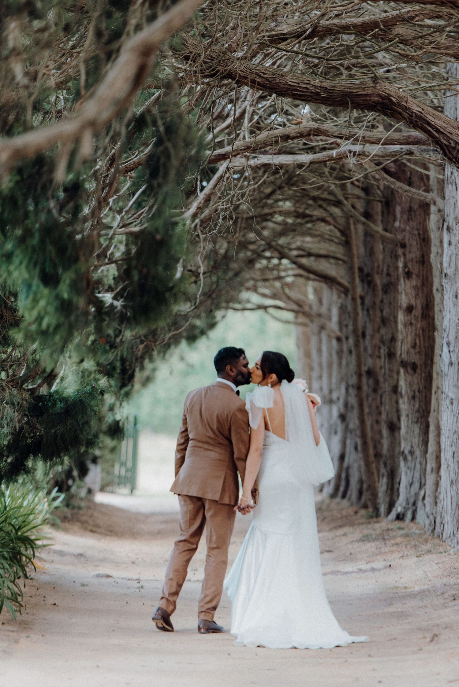 Couple standing on tree-lined path in elegant wedding attire.