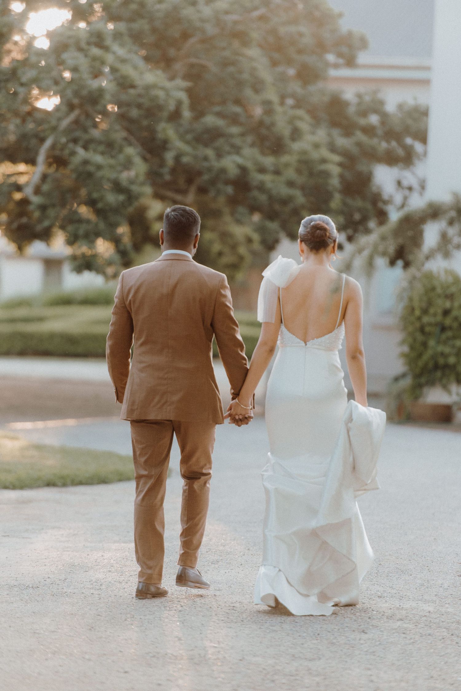 Couple in wedding attire walking hand in hand down a path at sunset.