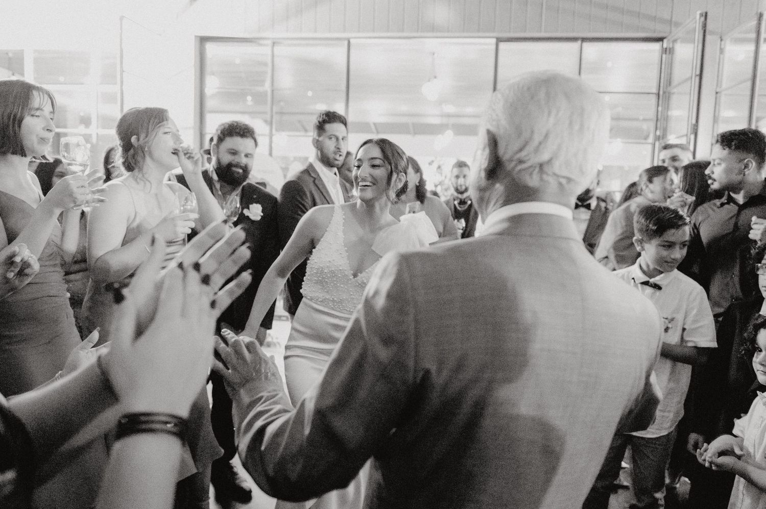 Wedding guests dance and celebrate in a bright reception hall captured in black and white.
