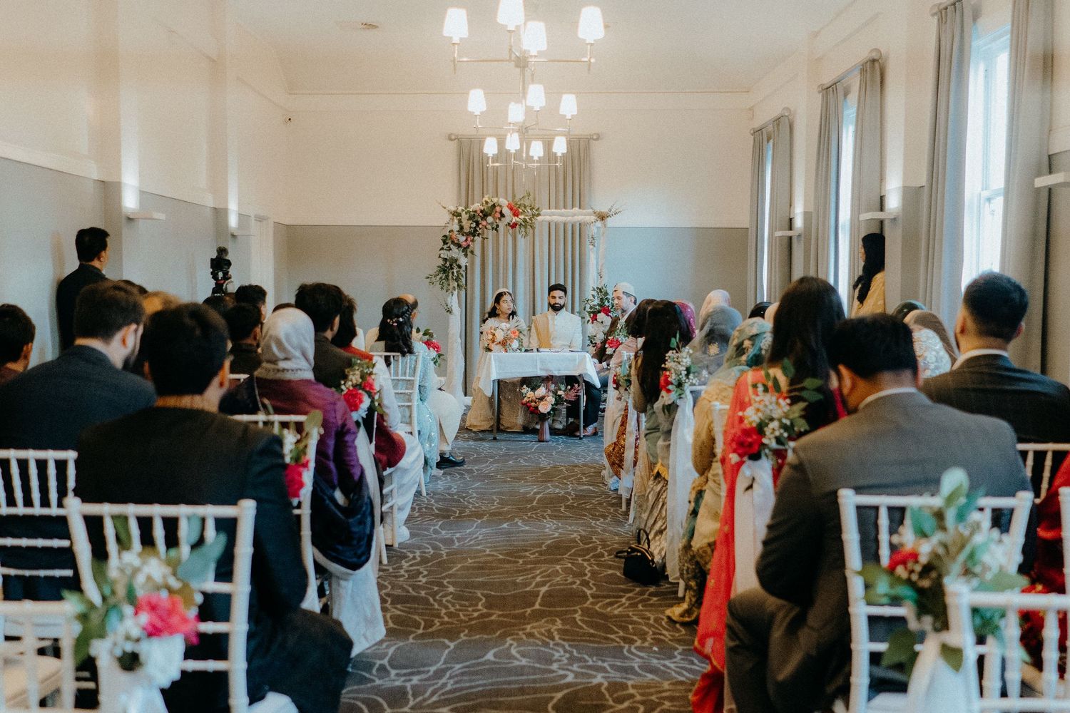 Wedding ceremony in progress with guests seated in white chairs in an elegant indoor venue.