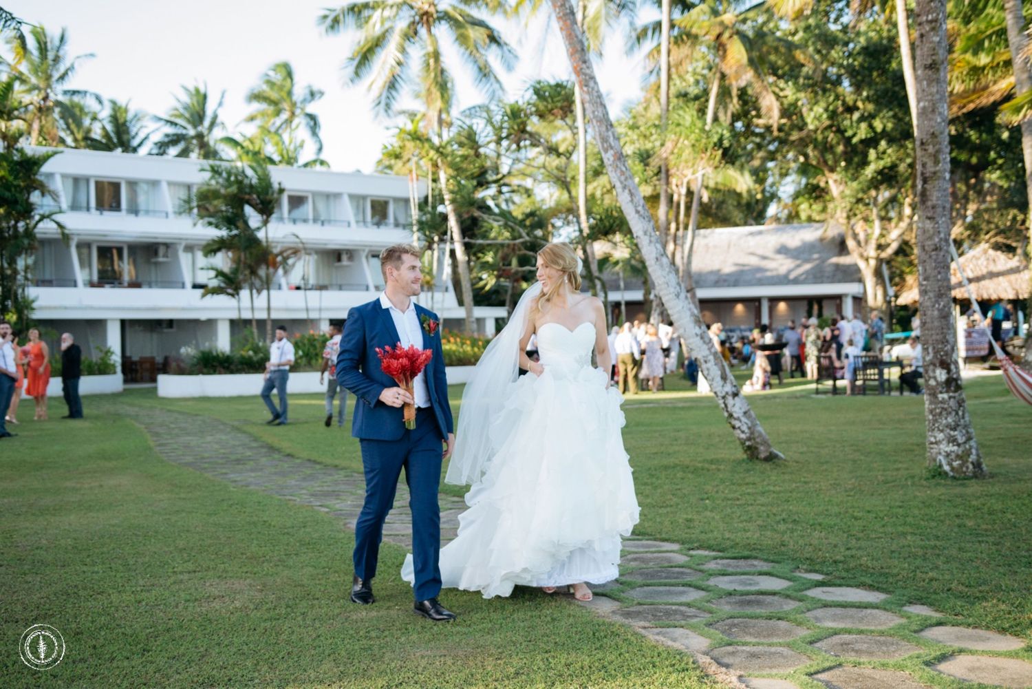 A couple walking on a path surrounded by palm trees and white buildings.