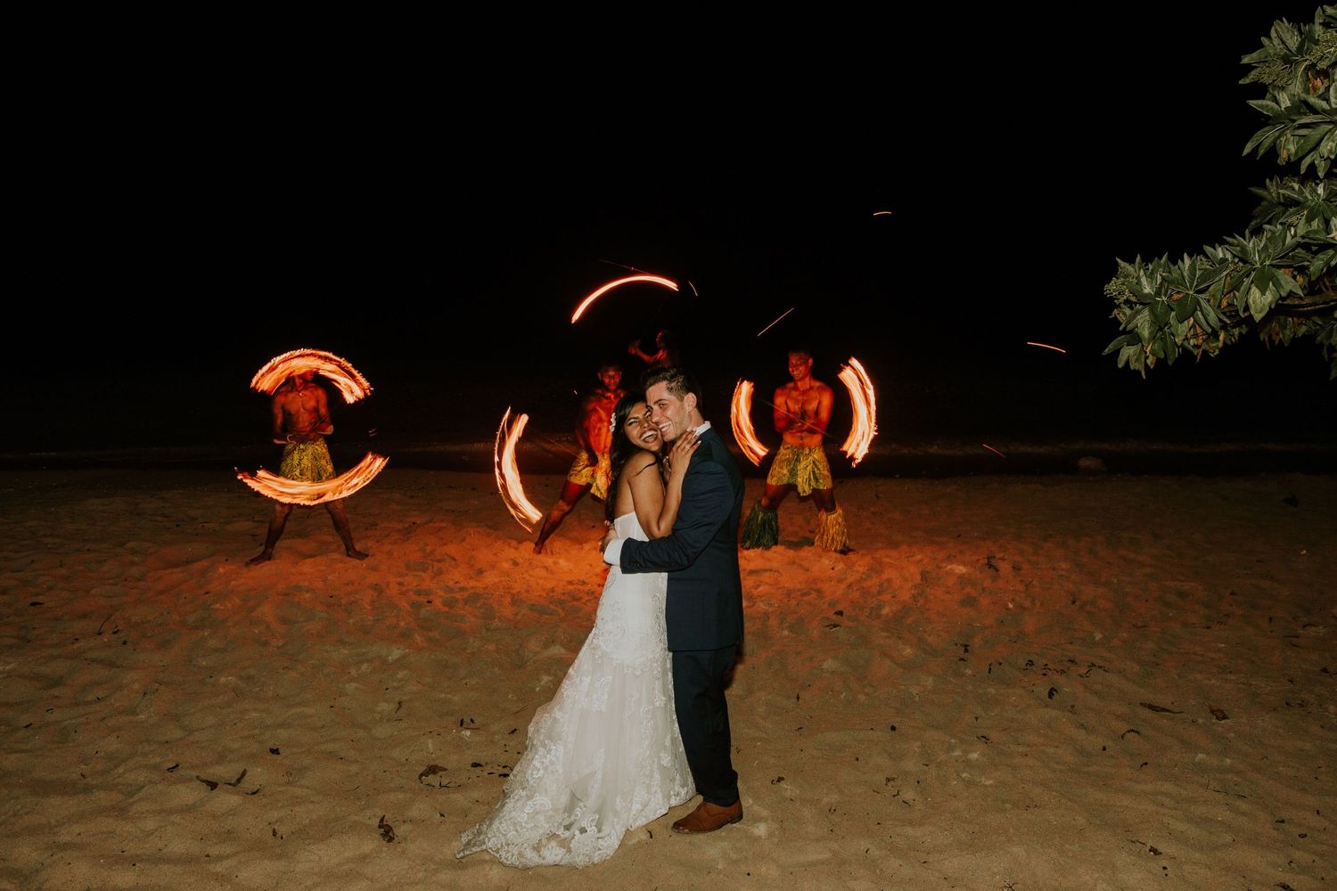 A couple dances on a beach at night while fire performers create circular light patterns in the background.