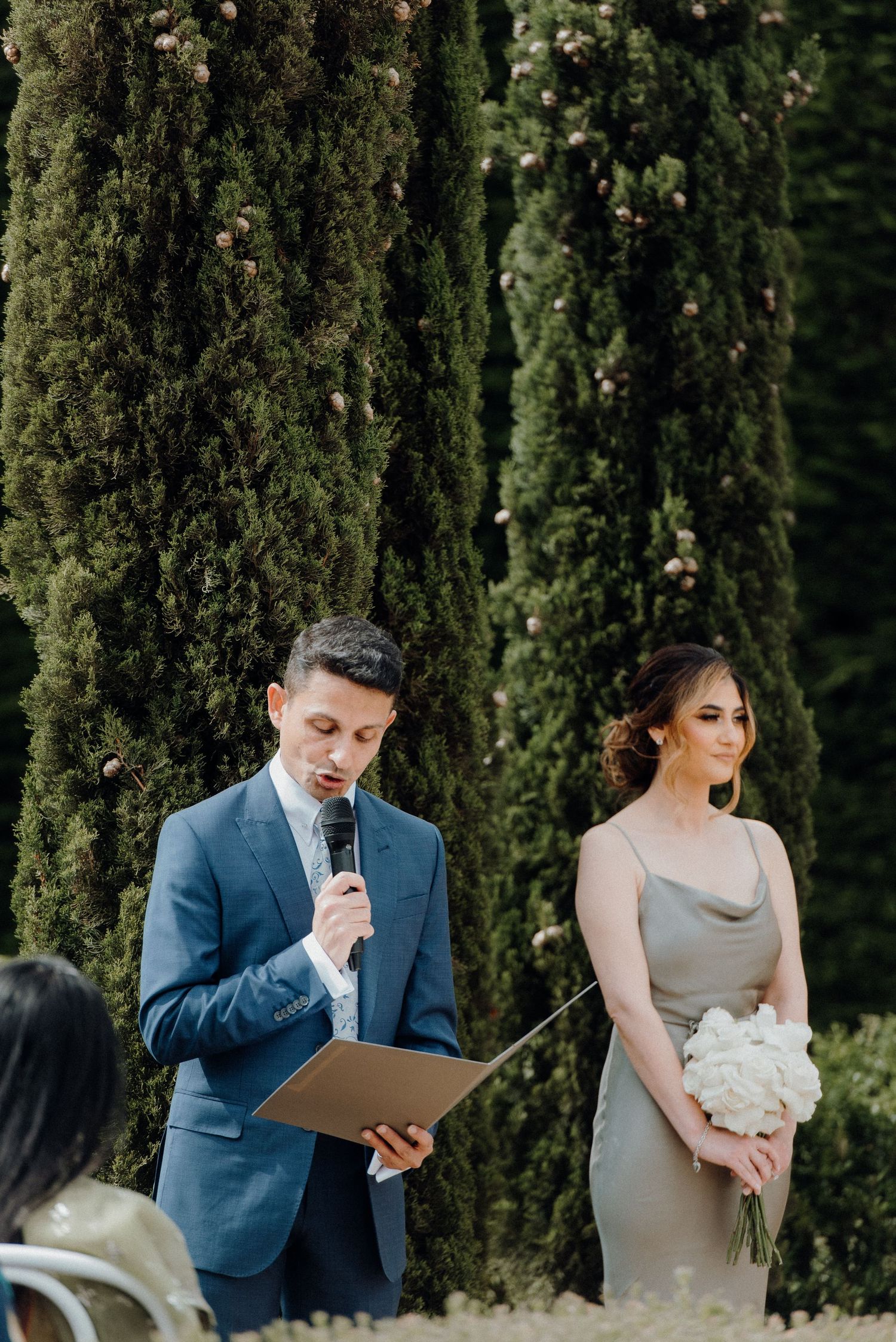 Wedding ceremony moment with speakers and attendants against a backdrop of cypress trees.
