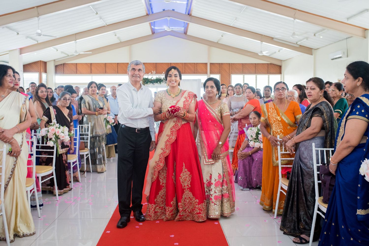 Couple walks down red carpet aisle while wedding guests applaud in bright reception hall.