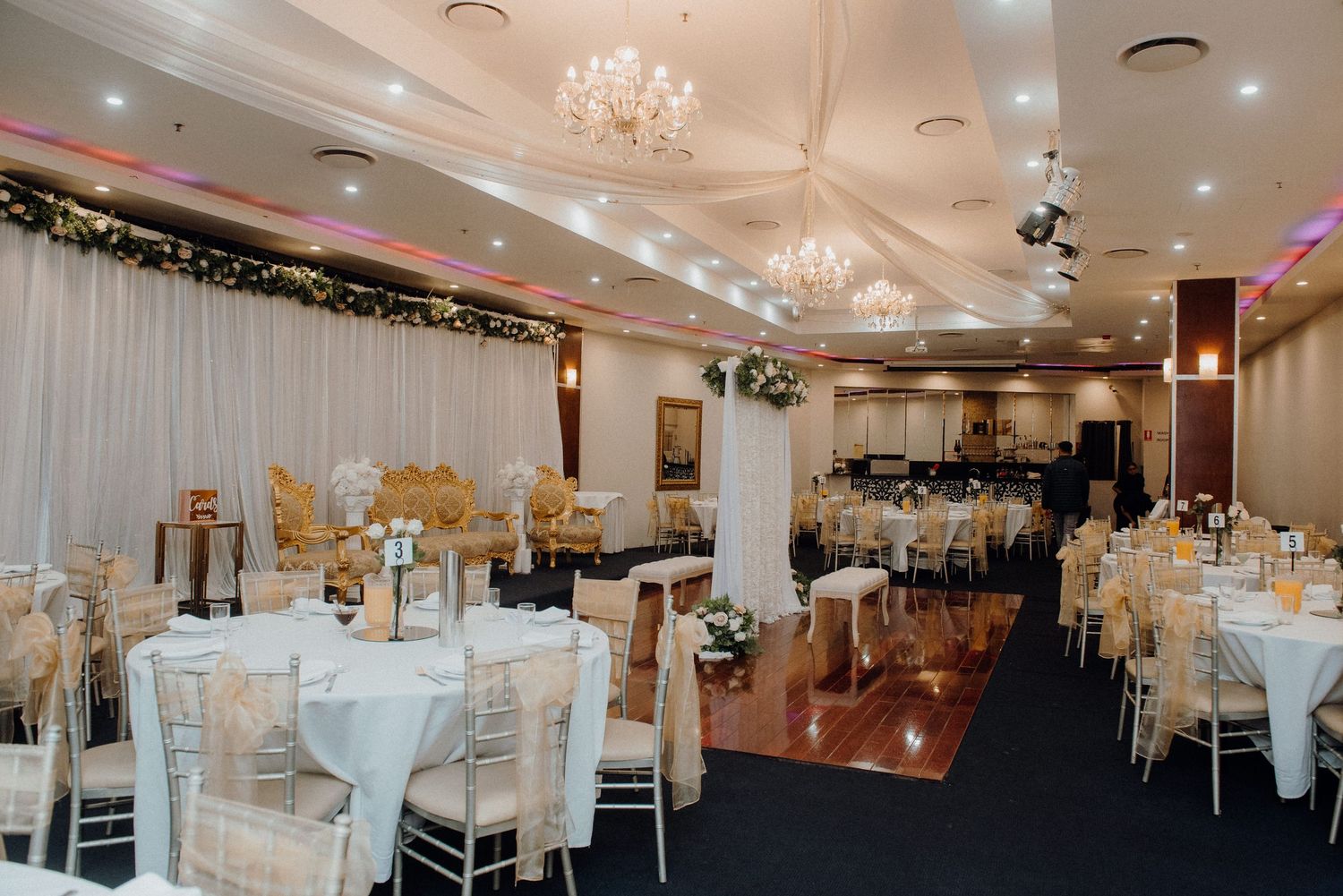 Elegant wedding reception venue with crystal chandeliers, white linens, and floral decorations along a draped wall.