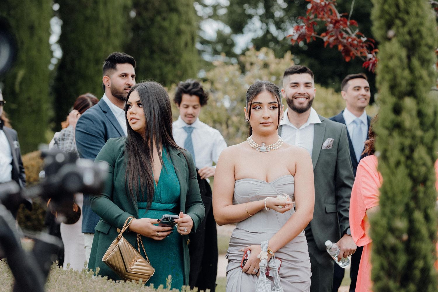 Wedding guests in formal attire gathered among tall cypress trees.