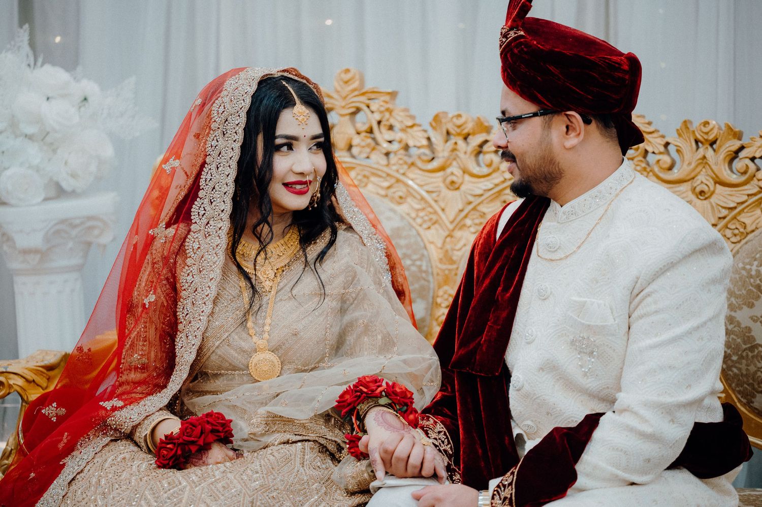 A couple in traditional South Asian wedding attire seated on an ornate golden throne during their ceremony.