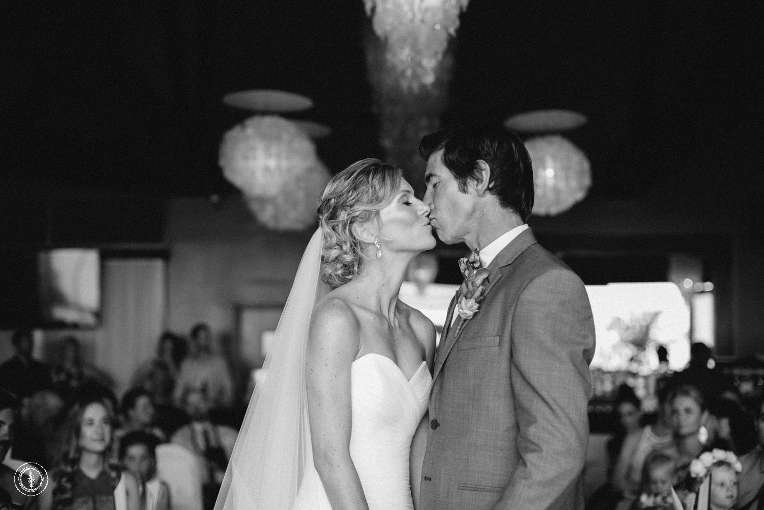 Bride and groom share their first kiss at the altar surrounded by wedding guests in black and white.