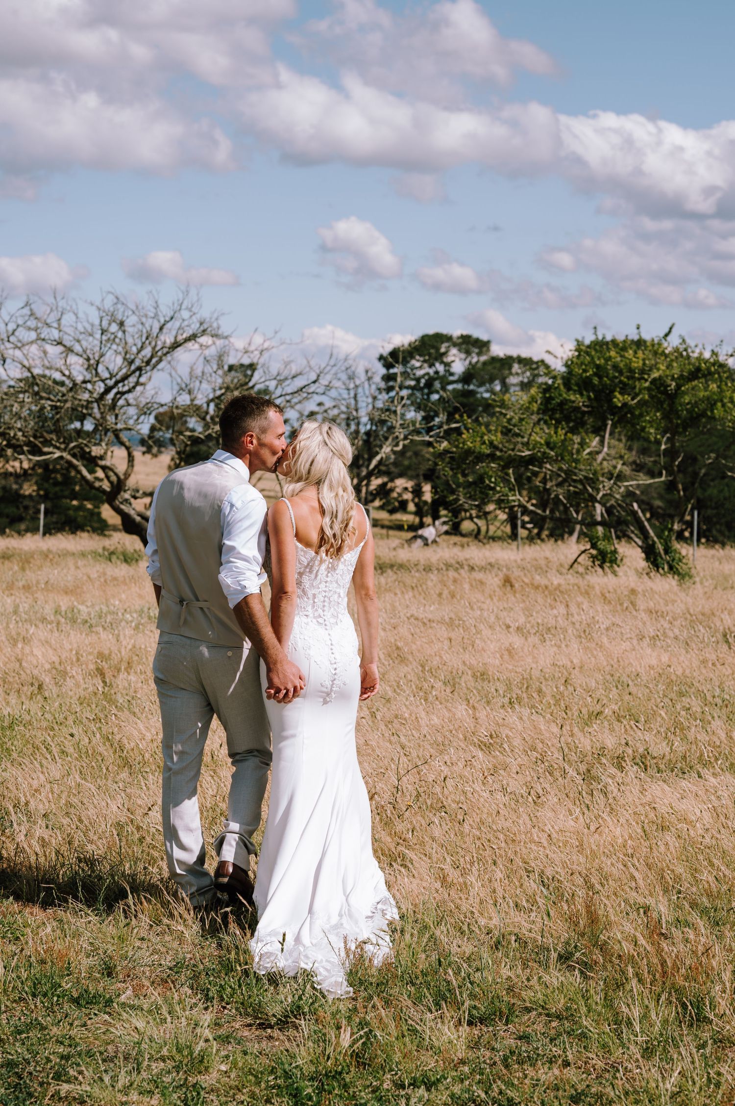 Bride and groom kiss while holding hands in a golden field under a blue sky on their wedding day.