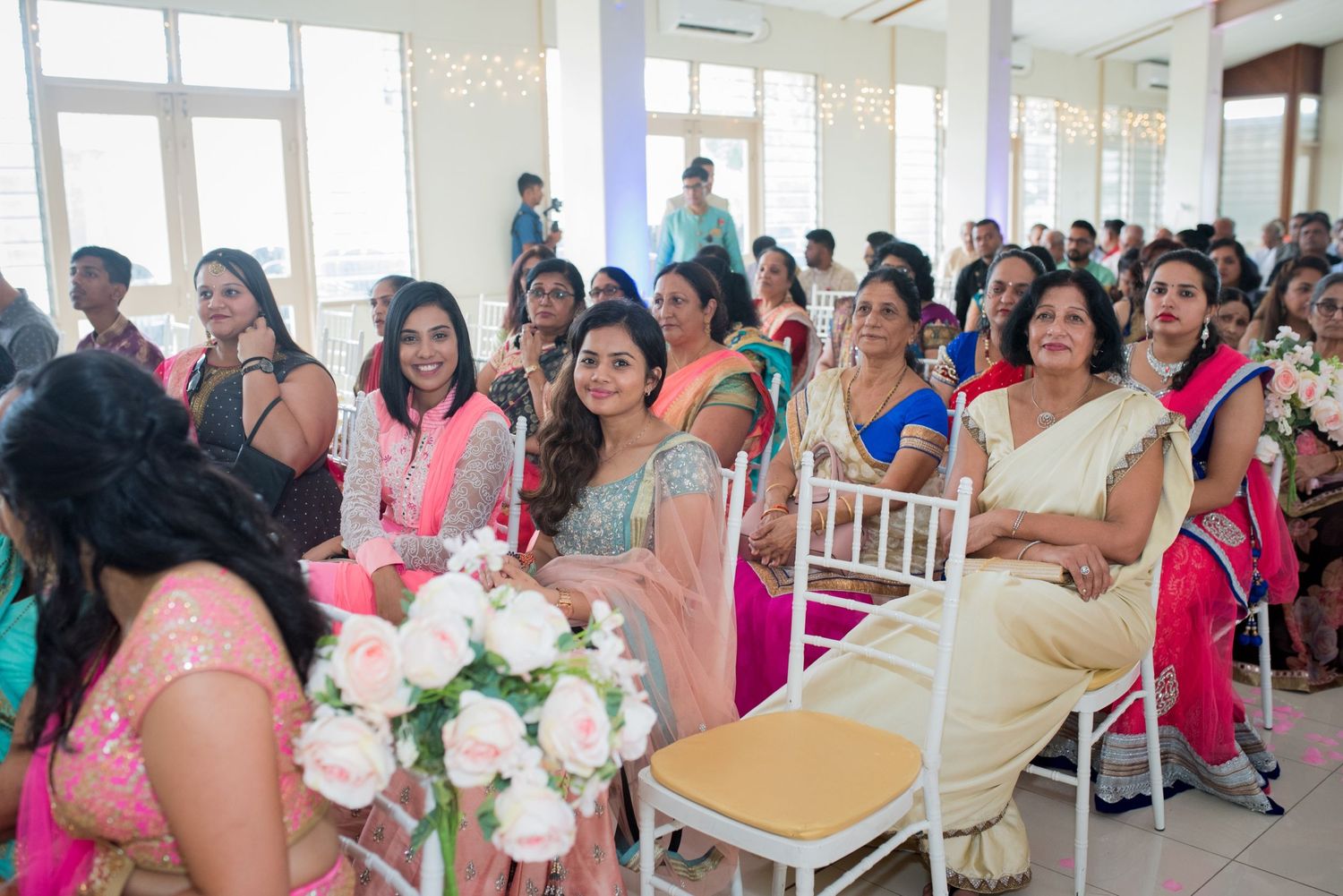 Wedding guests seated in white chairs attending a celebration in a bright indoor venue.
