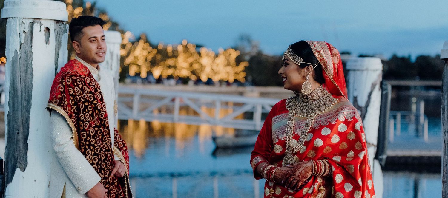 A couple in traditional South Asian wedding attire pose together at sunset by a waterfront.
