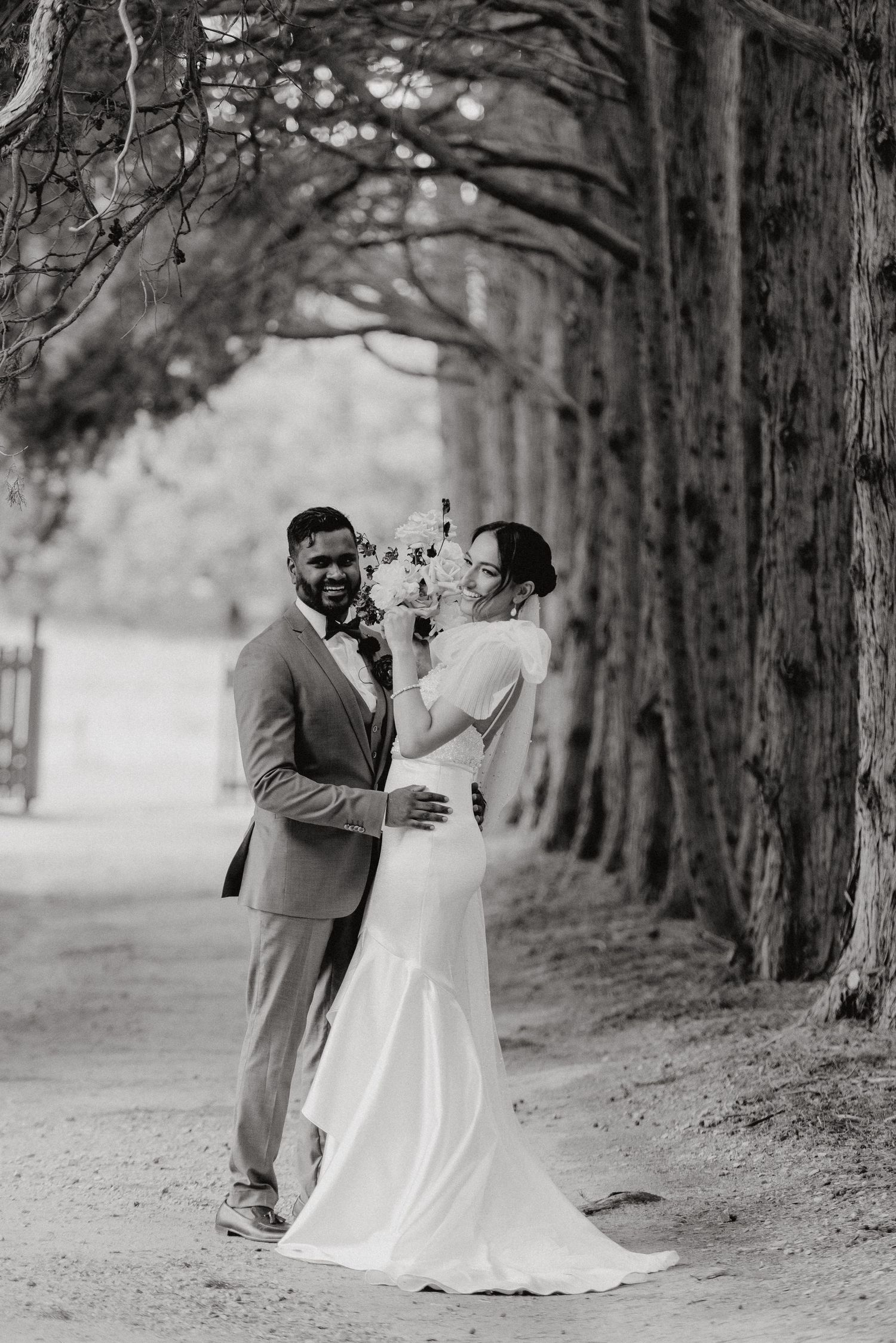 Black and white photo of couple embracing on tree-lined path.