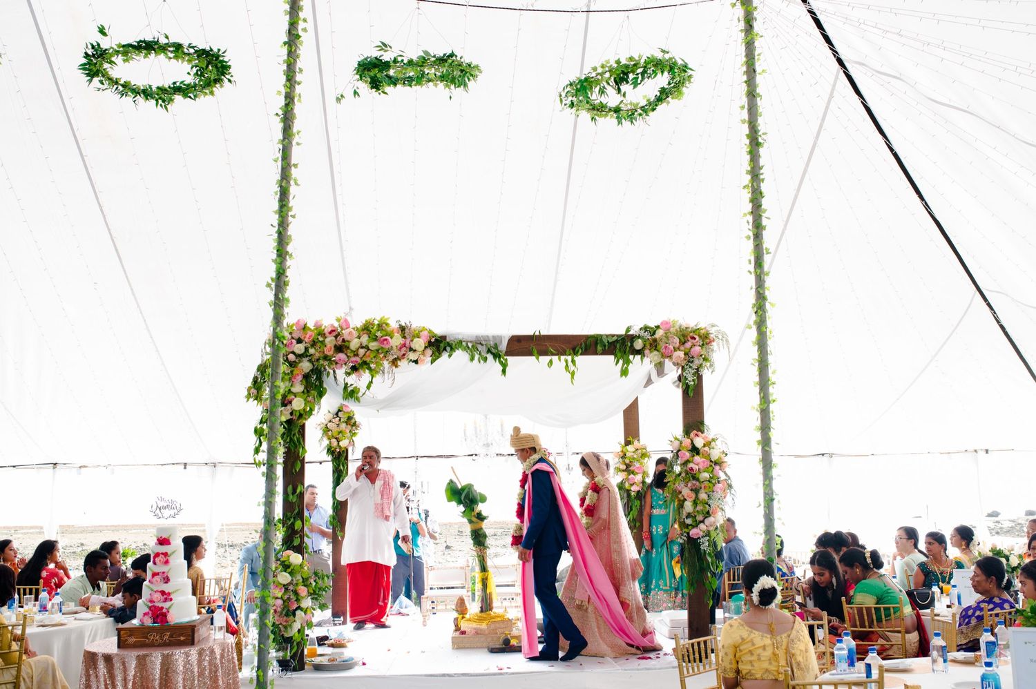 Wedding ceremony setup under a white tent with floral garlands and guests seated.