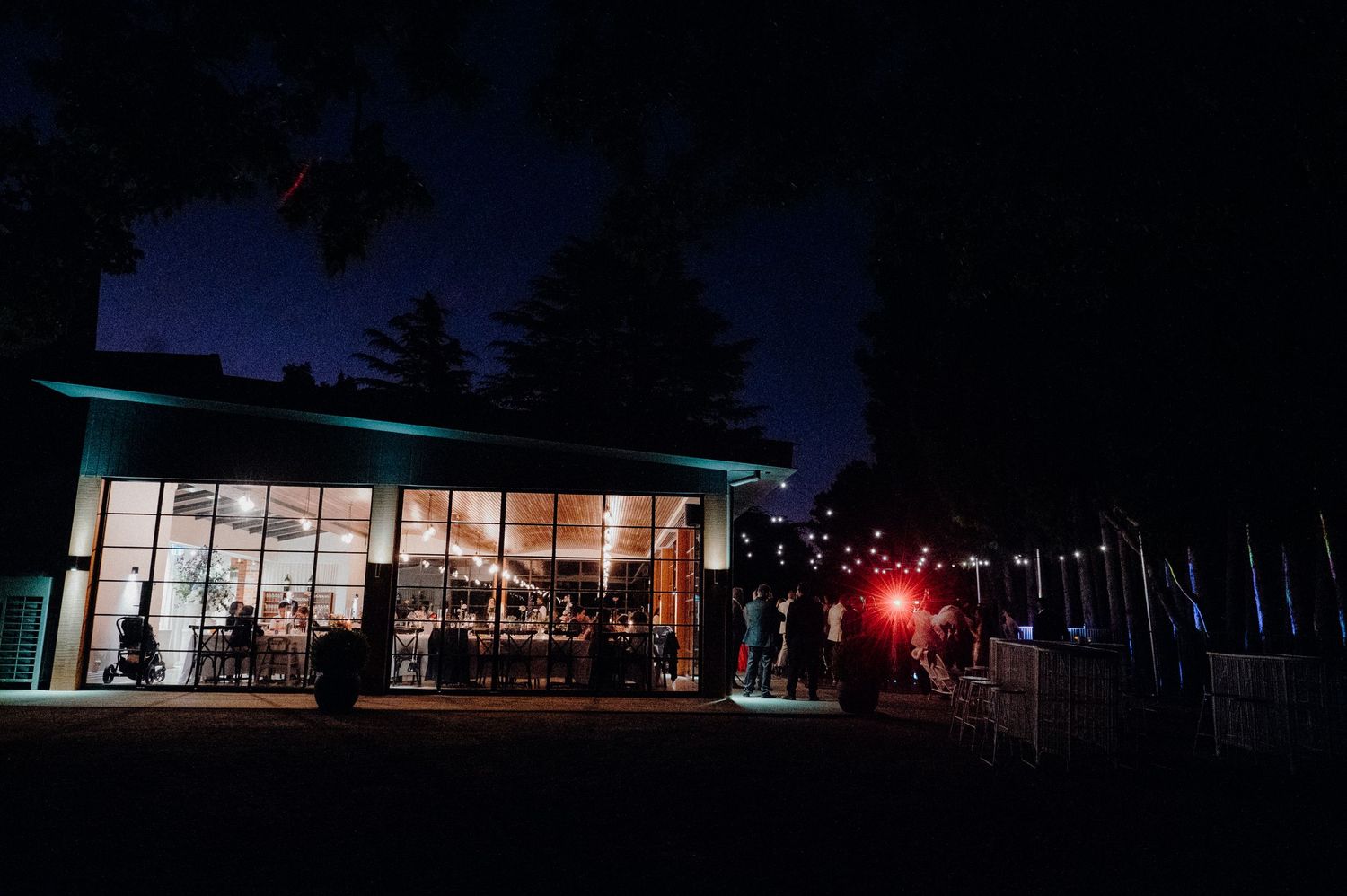 Modern glass building illuminated at night with string lights and wedding guests outside.