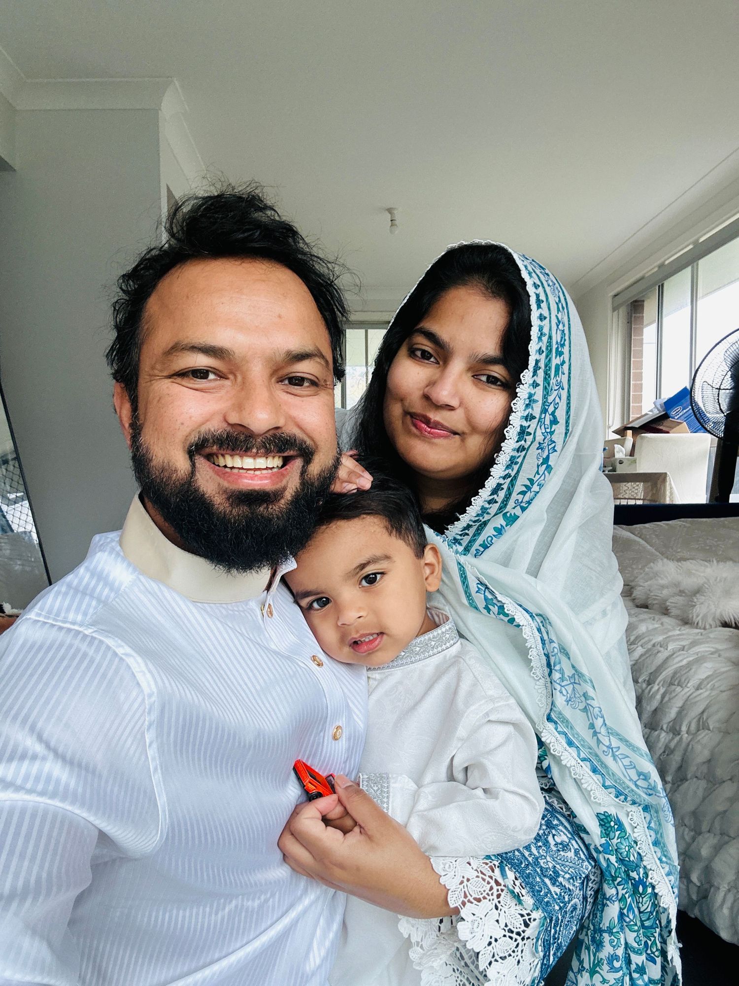 Happy South Asian family selfie with bearded father, mother in blue hijab holding baby, all dressed in white.