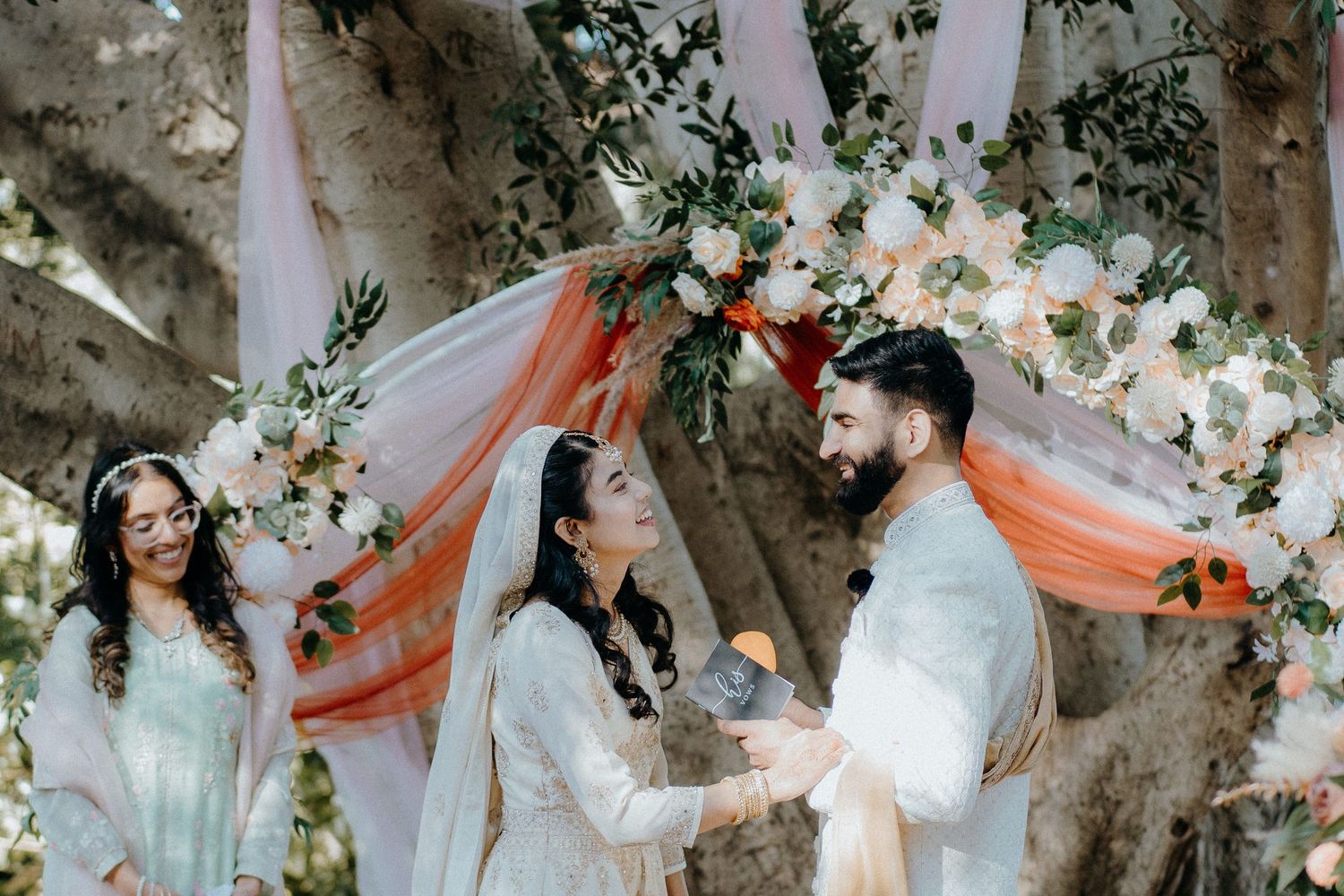 Wedding ceremony under a decorated floral arch with orange and white accents in an outdoor setting.