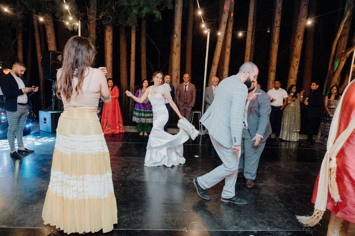 Wedding guests dancing at an outdoor reception with string lights and forest scenery.