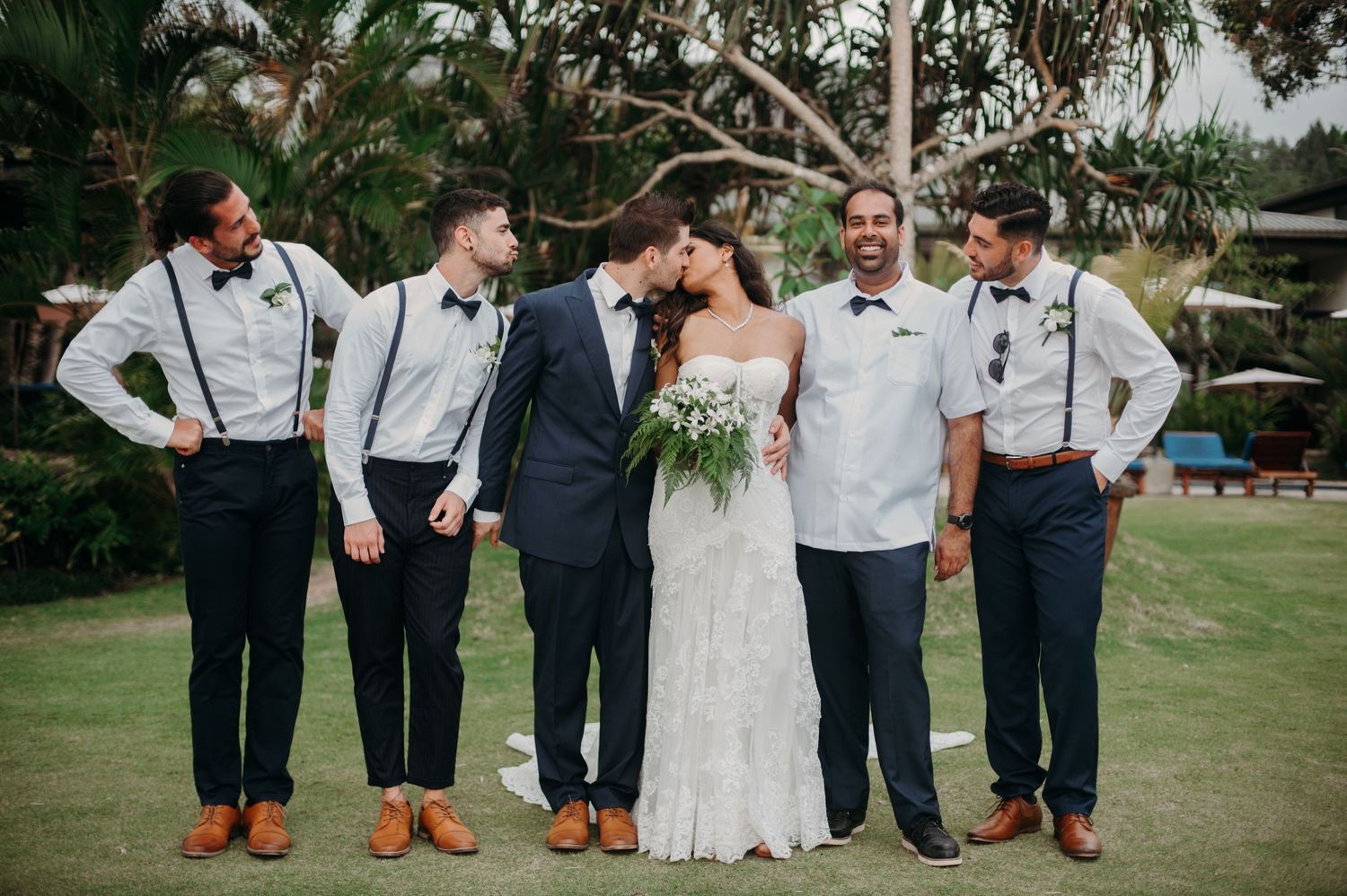 Wedding party in formal attire with suspenders and bow ties posing on green lawn.