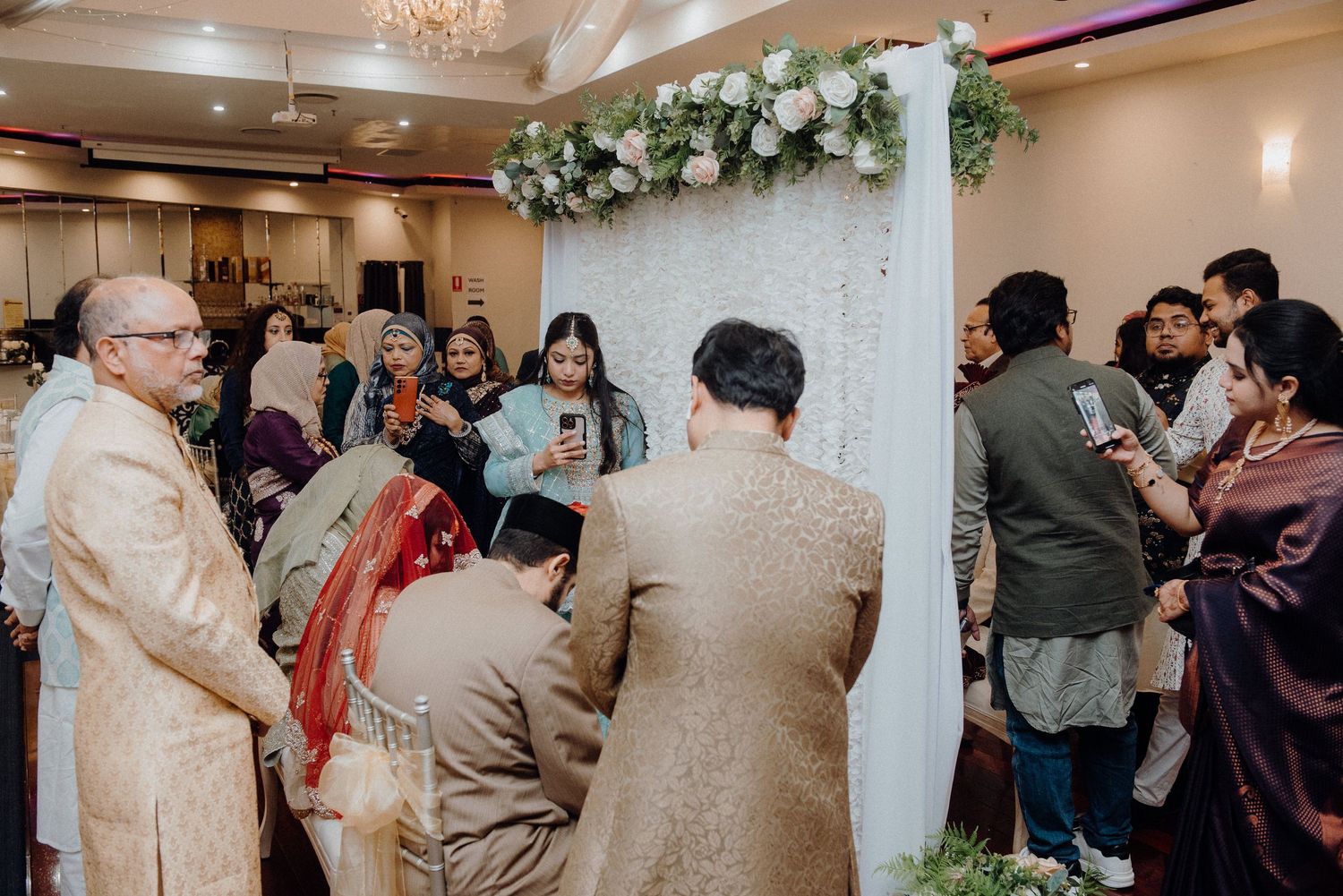 Guests gather around an ornate white pillar decorated with floral arrangements at a traditional South Asian wedding ceremony.