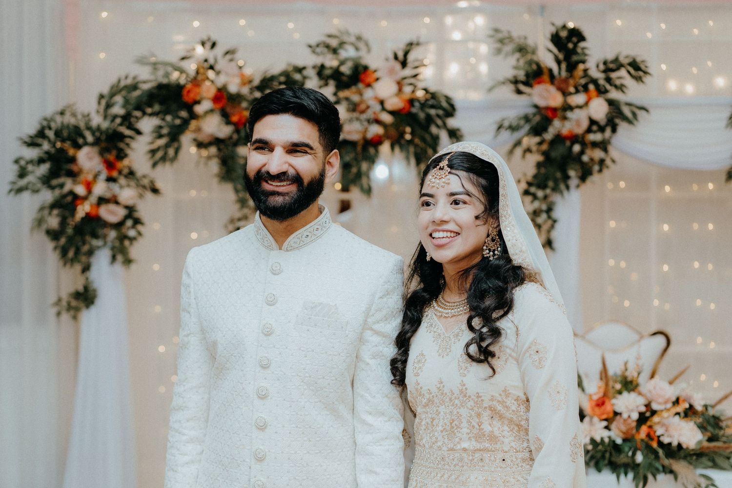 A wedding couple in traditional white attire poses against a backdrop of flowers and string lights.