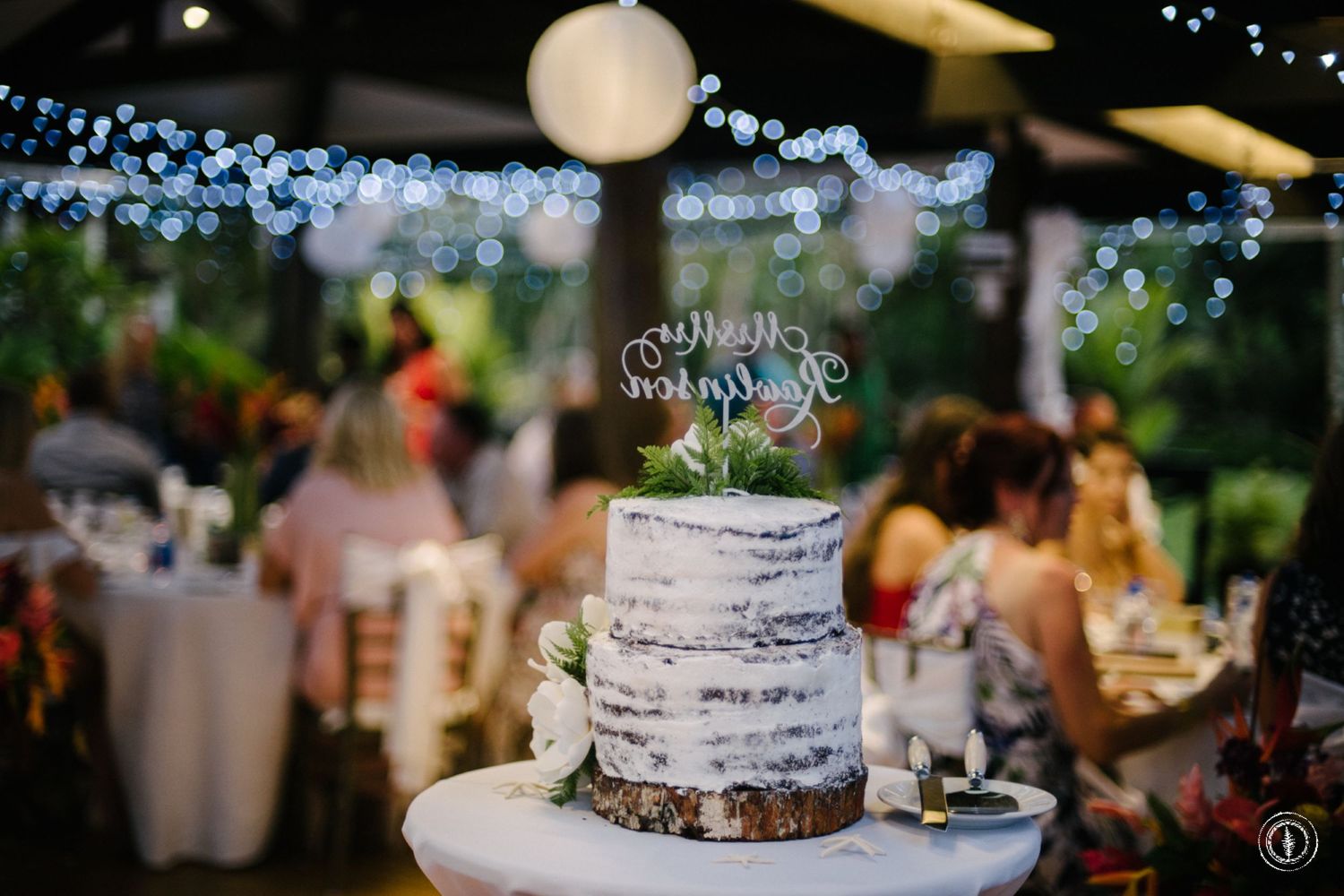 Rustic white wedding cake displayed under twinkle lights at an outdoor reception.
