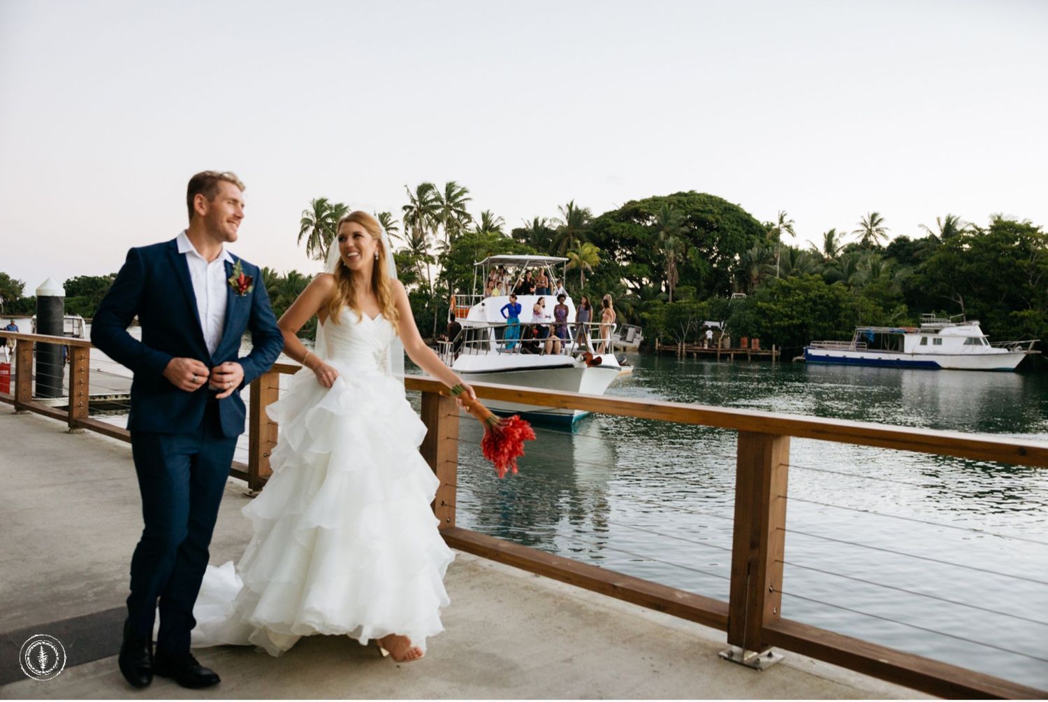 Wedding couple strolls along marina boardwalk with boats and palm trees in background.