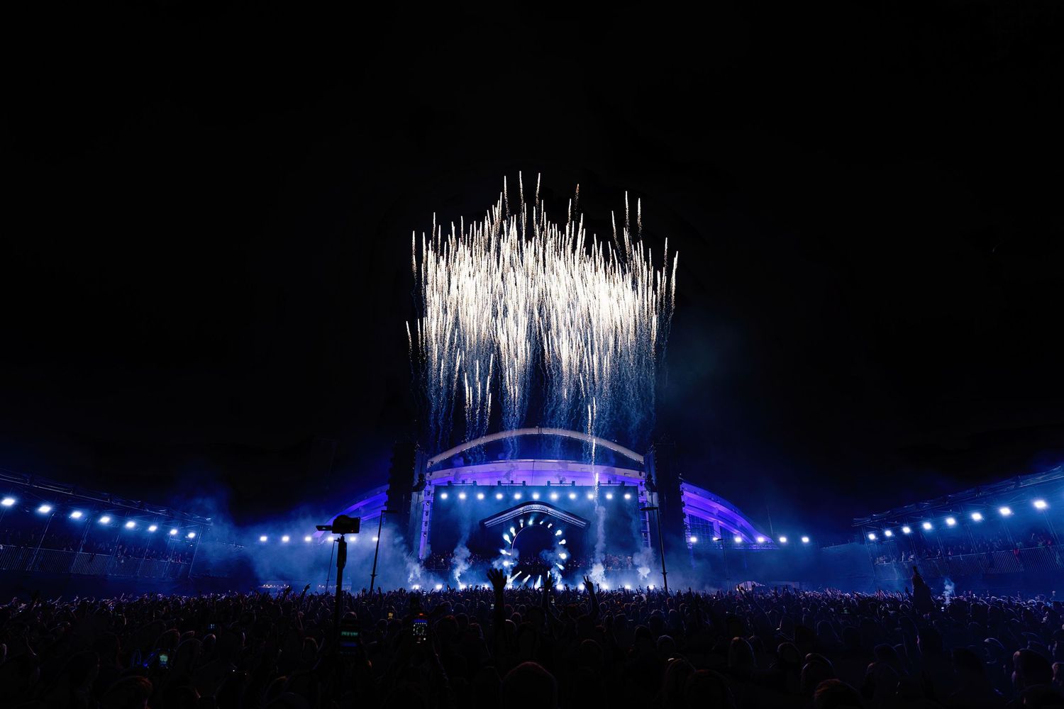 Spectacular fireworks explode above a blue-lit concert stage as thousands of fans watch a live outdoor music festival. Armin van Buuren.