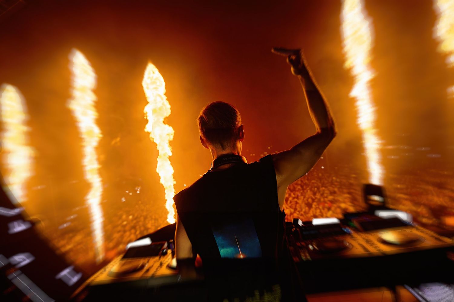 DJ Armin van Buuren performing on stage with big flames and crowd in background at a music festival.