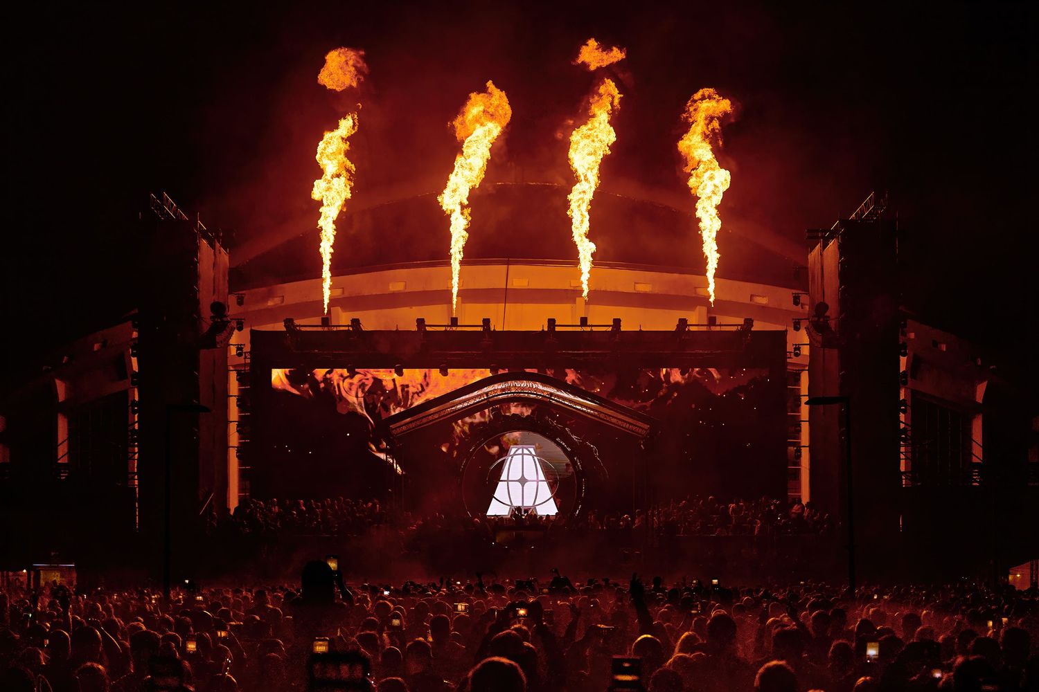 Massive outdoor music festival stage with four towering flame cannons firing against a dark night sky. Armin van Buuren.