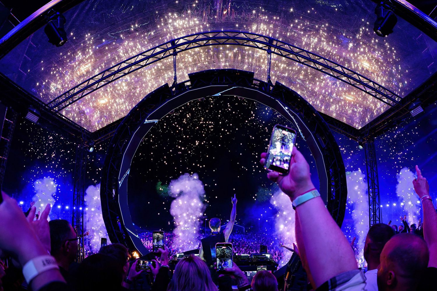 Crowd at a vibrant music festival with pyrotechnical jets, colorful lights, and a glowing stage arch overhead.