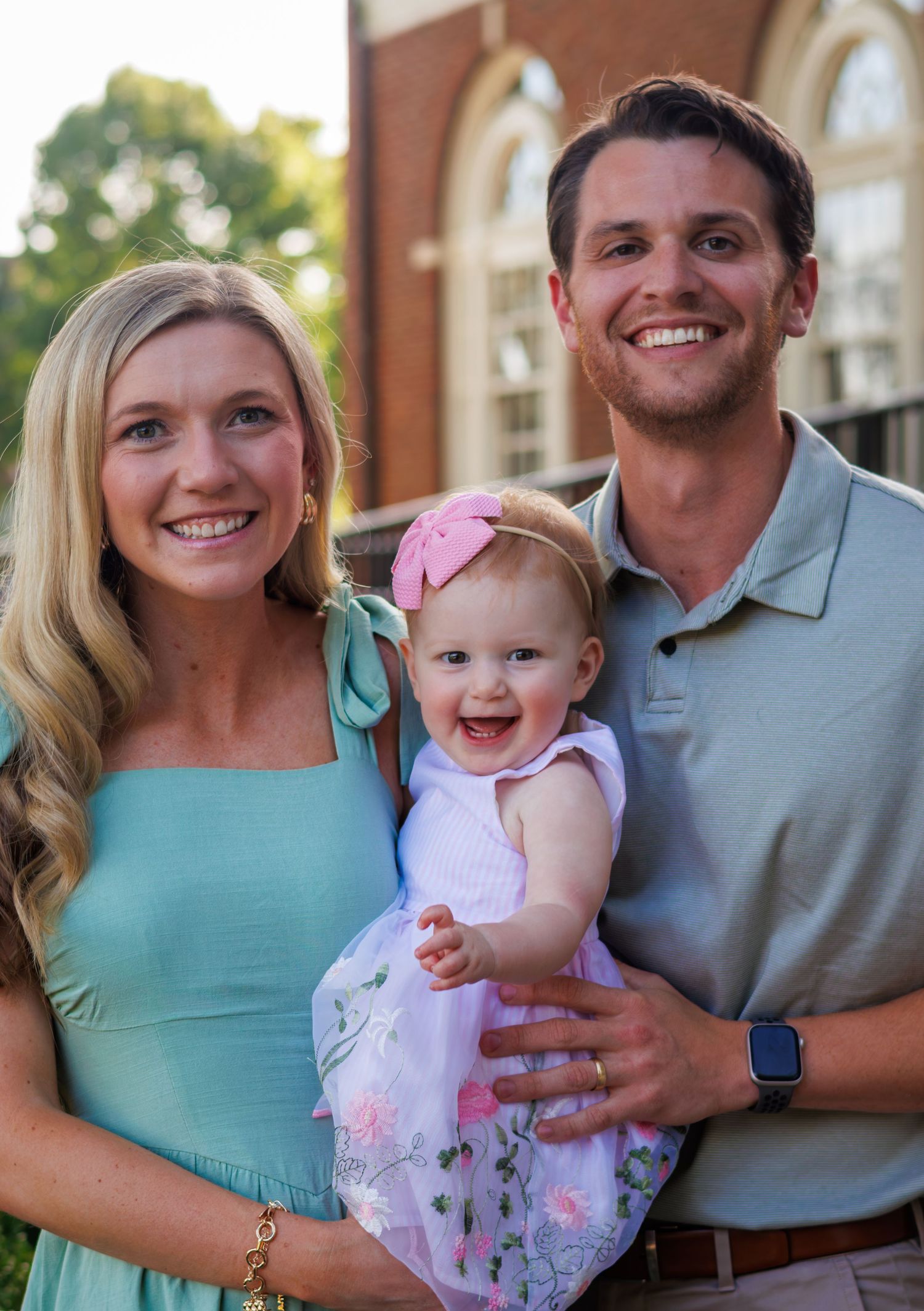 Happy young family with smiling parents holding their baby girl in pink dress outdoors near brick building.