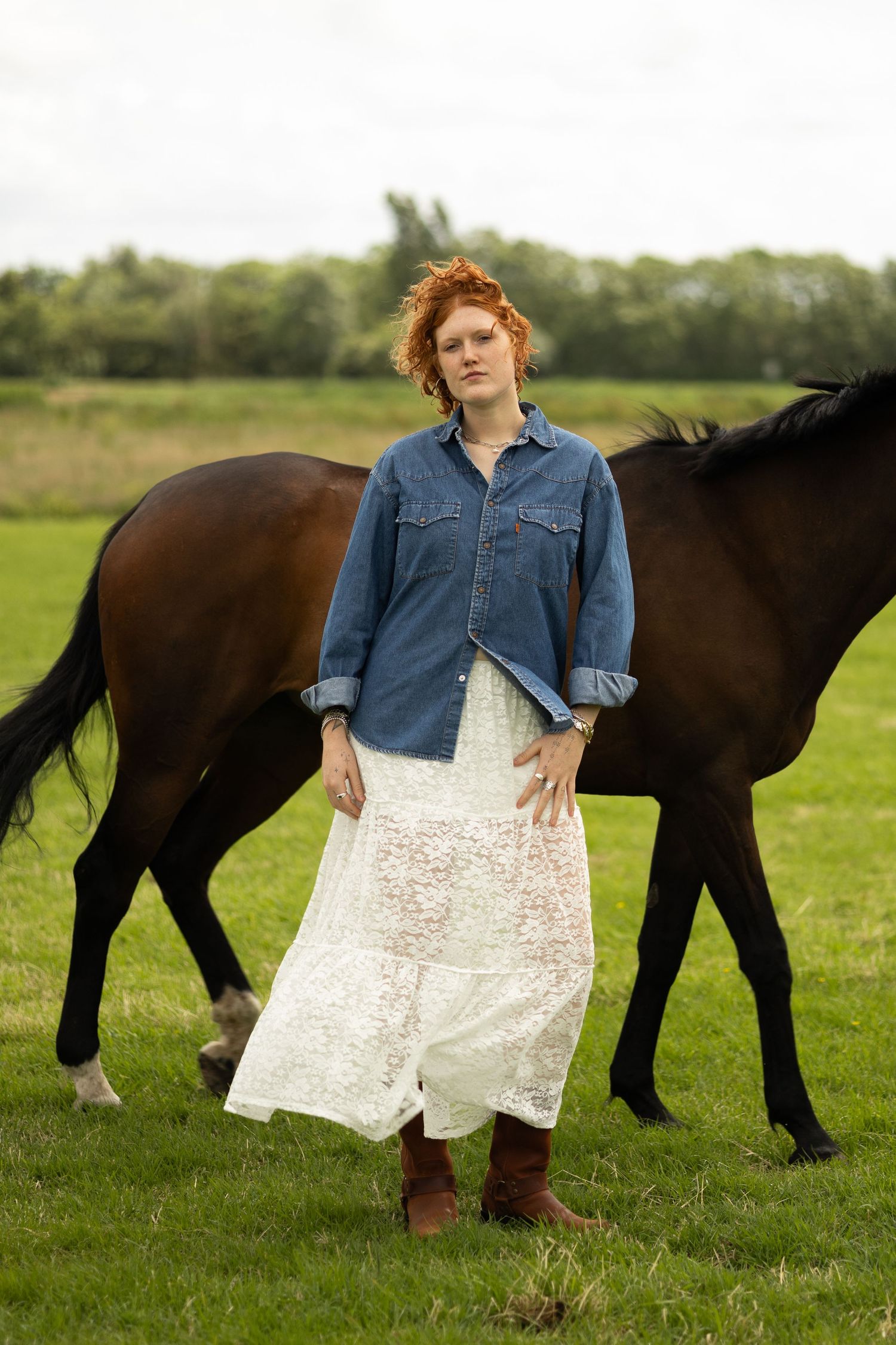 Red-haired woman in denim jacket and white lace skirt standing in a field with a brown horse behind her.