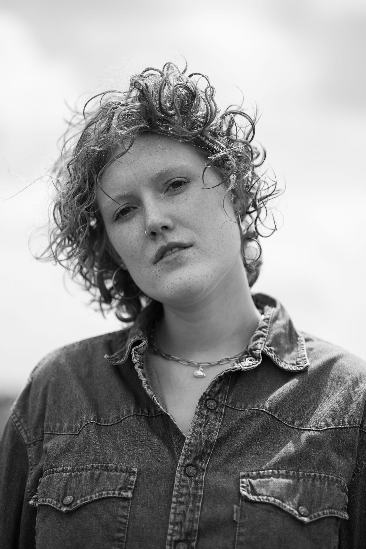 Black and white portrait of a curly-haired woman in a denim shirt wearing a delicate necklace outdoors.
