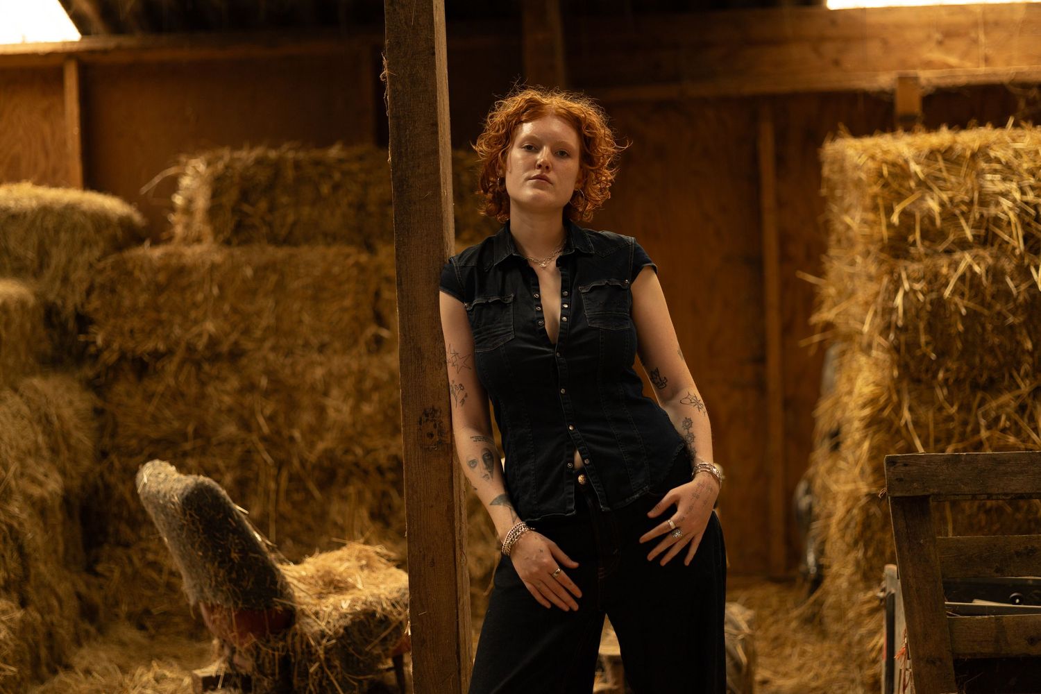 Red-haired tattooed woman in black jumpsuit posing confidently in a barn surrounded by hay bales.